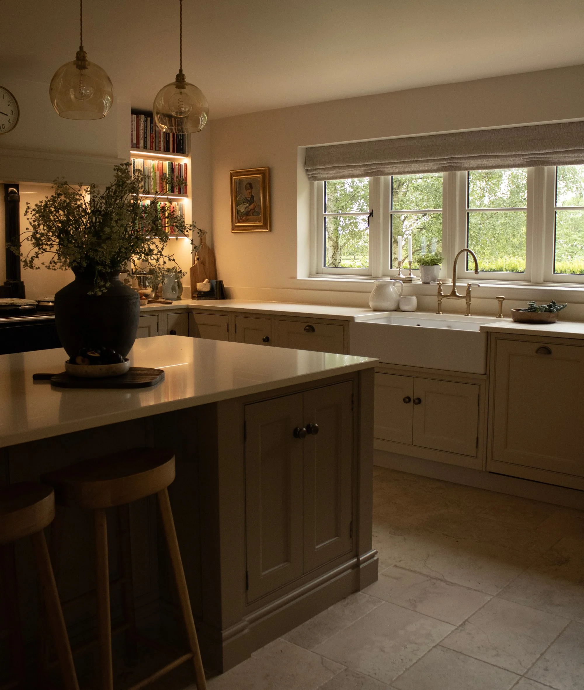 Kitchen with large window, beige cabinetry, white countertop, and potted plants. Wooden chairs are paired at the kitchen island, and hanging glass light fixtures are above. A framed painting is on the wall, and books are on a shelf near the light.