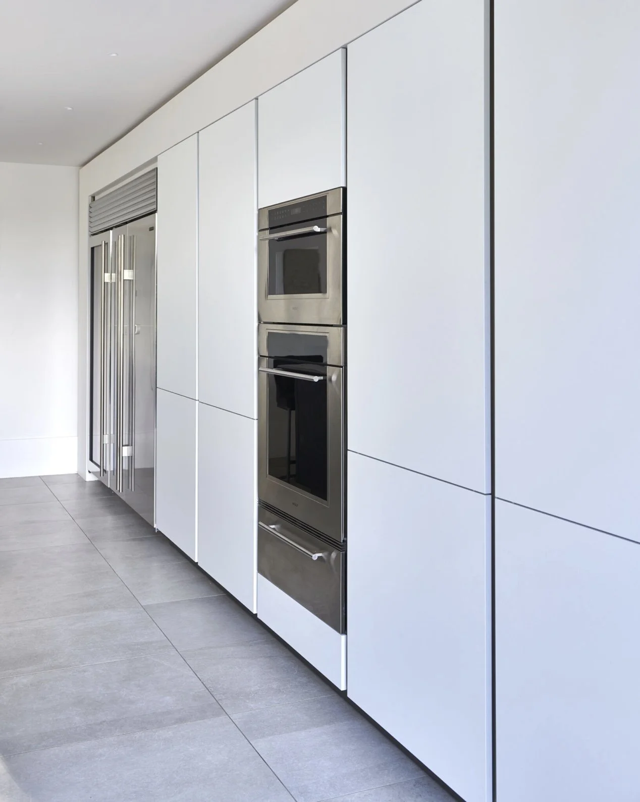 Modern kitchen with built-in stainless steel oven and microwave, white cabinets, gray tiled floor, and a refrigerator with sliding doors.