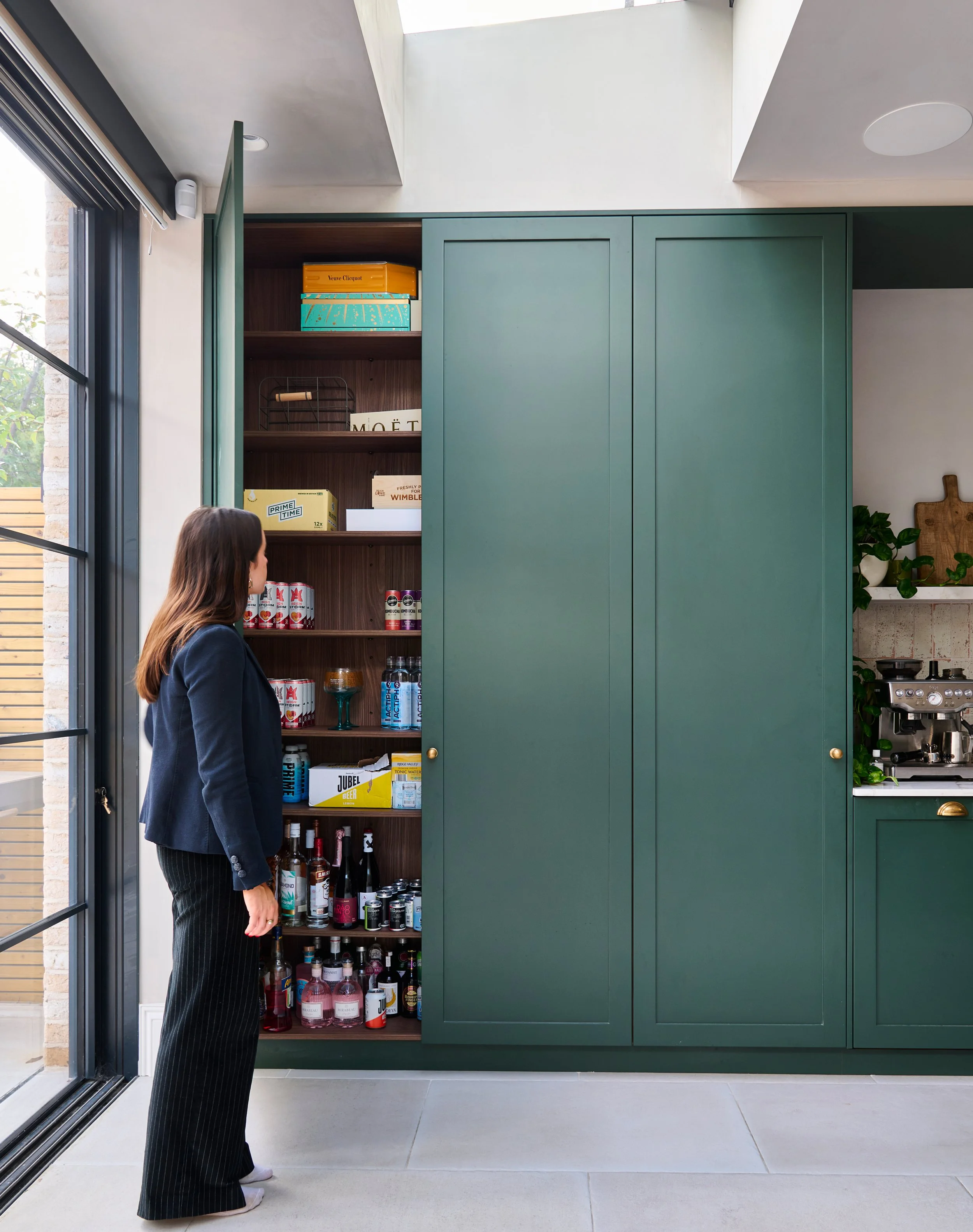 A woman with long brown hair wearing a dark blazer and pinstripe pants stands inside a modern kitchen, looking at a tall green cabinet with sliding doors and open shelving filled with various bottles, boxes, and cans, near a large window and sliding 