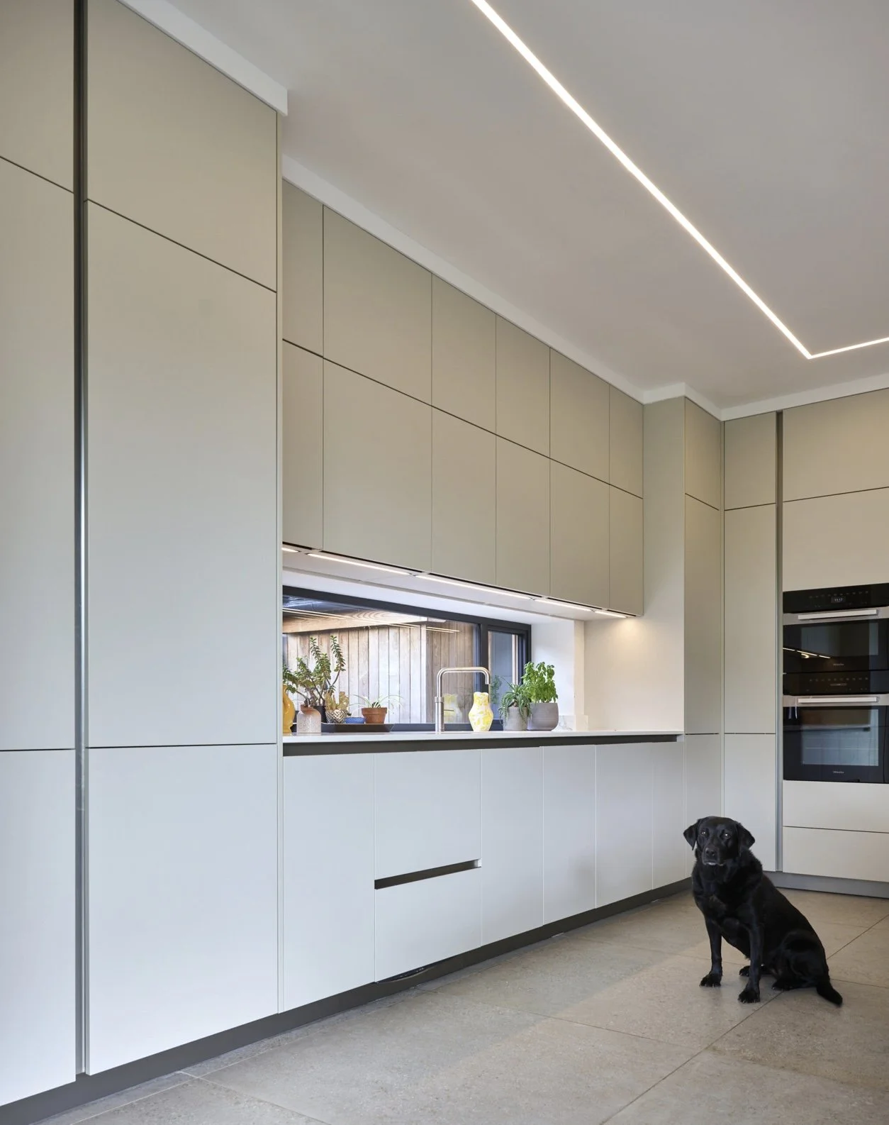 Modern kitchen with beige cabinetry, a small window with potted plants, and a black dog sitting on the floor.