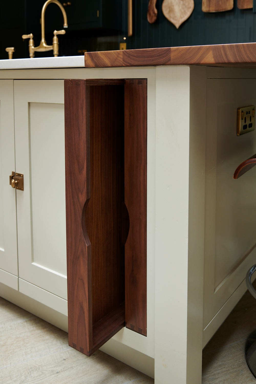 Close-up of a kitchen island with a drawer made of dark wood, partially pulled out, showing a unique oval-shaped cutout. The island has a white base, a wooden countertop, and a kitchen sink with a gold finish faucet in the background.