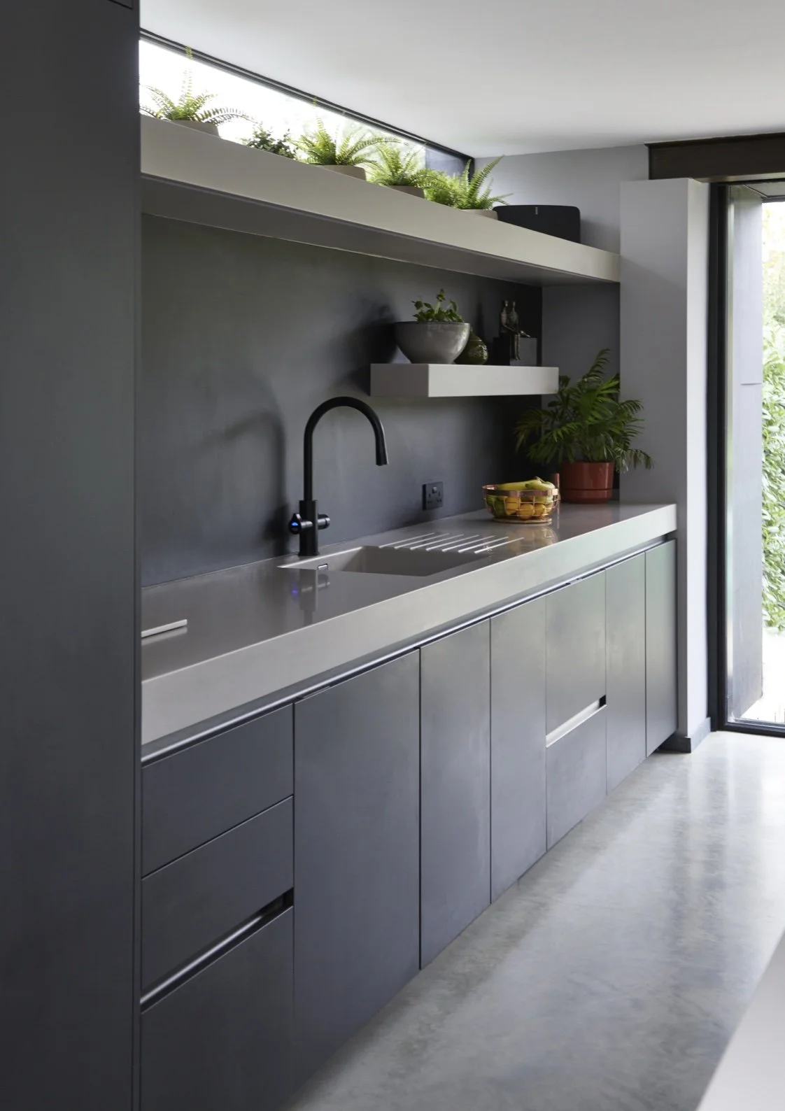 Modern kitchen with gray cabinets, black faucet, and plants on white shelves, with a sliding glass door open to greenery outside.
