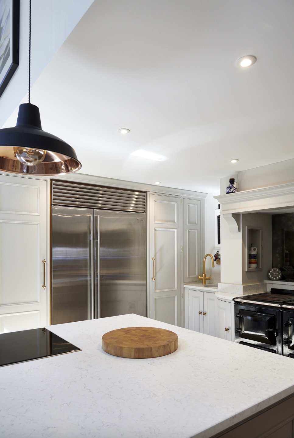 Modern kitchen with white cabinets, stainless steel refrigerator, black stove, marble countertop, wooden cutting board, gold faucet, and black and copper pendant light.