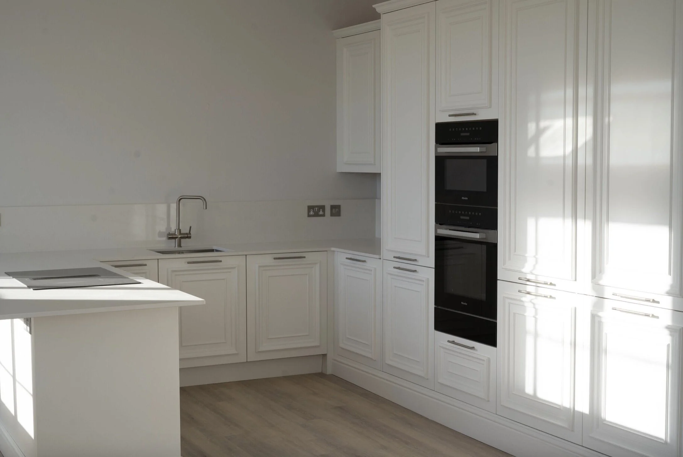 Kitchen with white cabinets, built-in black ovens, a sink with a chrome faucet, and light-colored wood flooring.