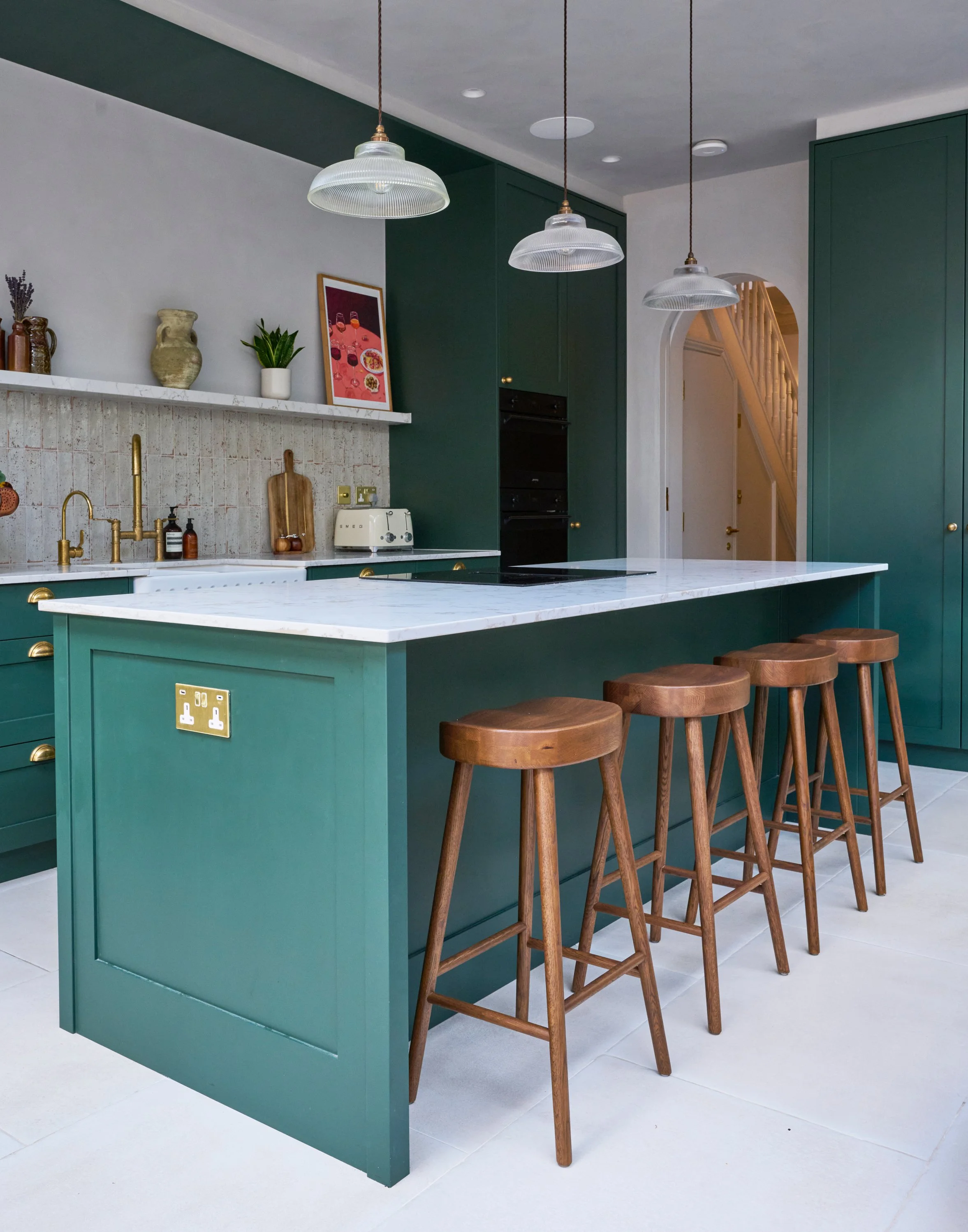 A modern kitchen with an island painted in teal, featuring a white marble countertop, and surrounded by four wooden bar stools. The backsplash is tiled with small beige tiles, and above it is a white shelf with decorative items including a vase, a pl