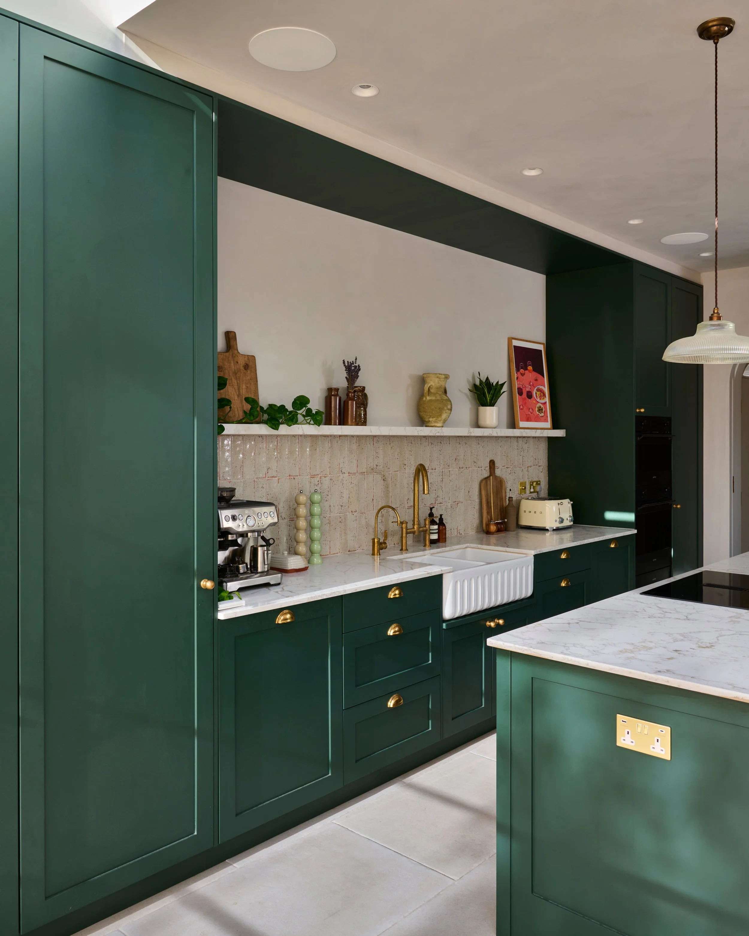 Green kitchen cabinets with gold handles, marble countertops, porcelain farmhouse sink, gold faucet, potted plant, small framed art, vintage bottles, coffee machine, toaster, and electrical outlets, with a white-tiled backsplash and ceiling lights.