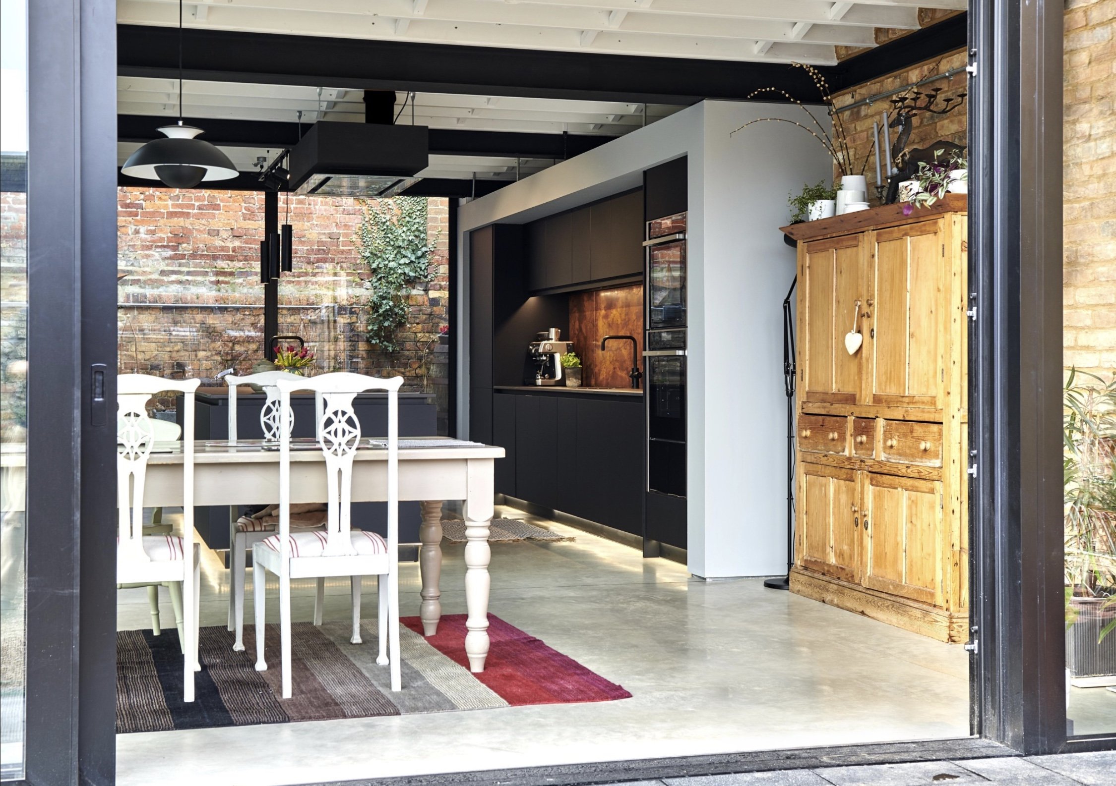 Modern kitchen and dining area with black cabinets, a white table, white chairs, and a rustic wooden cabinet, set in an open space with exposed brick walls and greenery visible outside.