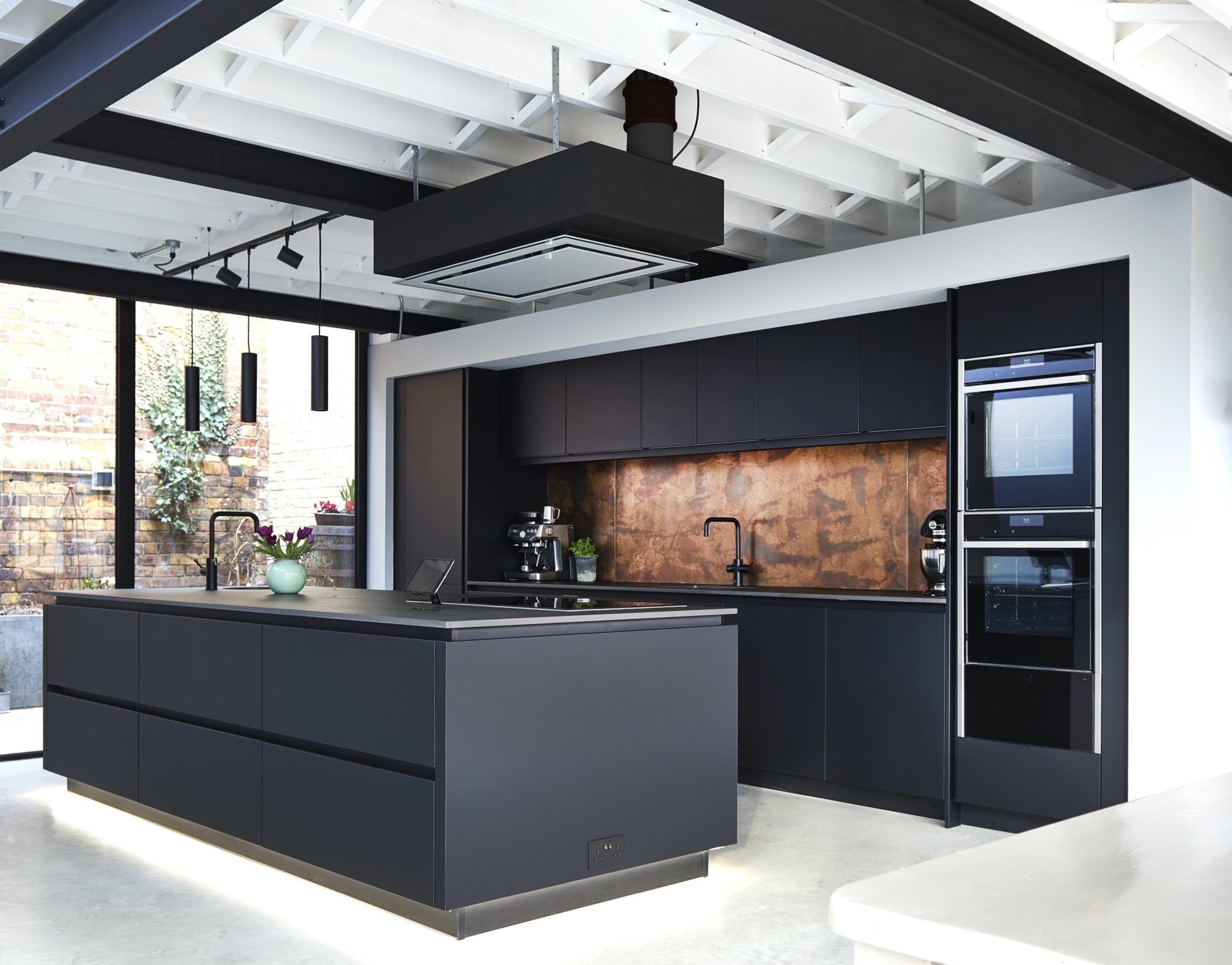 Modern kitchen with black cabinetry, copper backsplash, and large island with a vase of purple flowers and kitchen appliances.