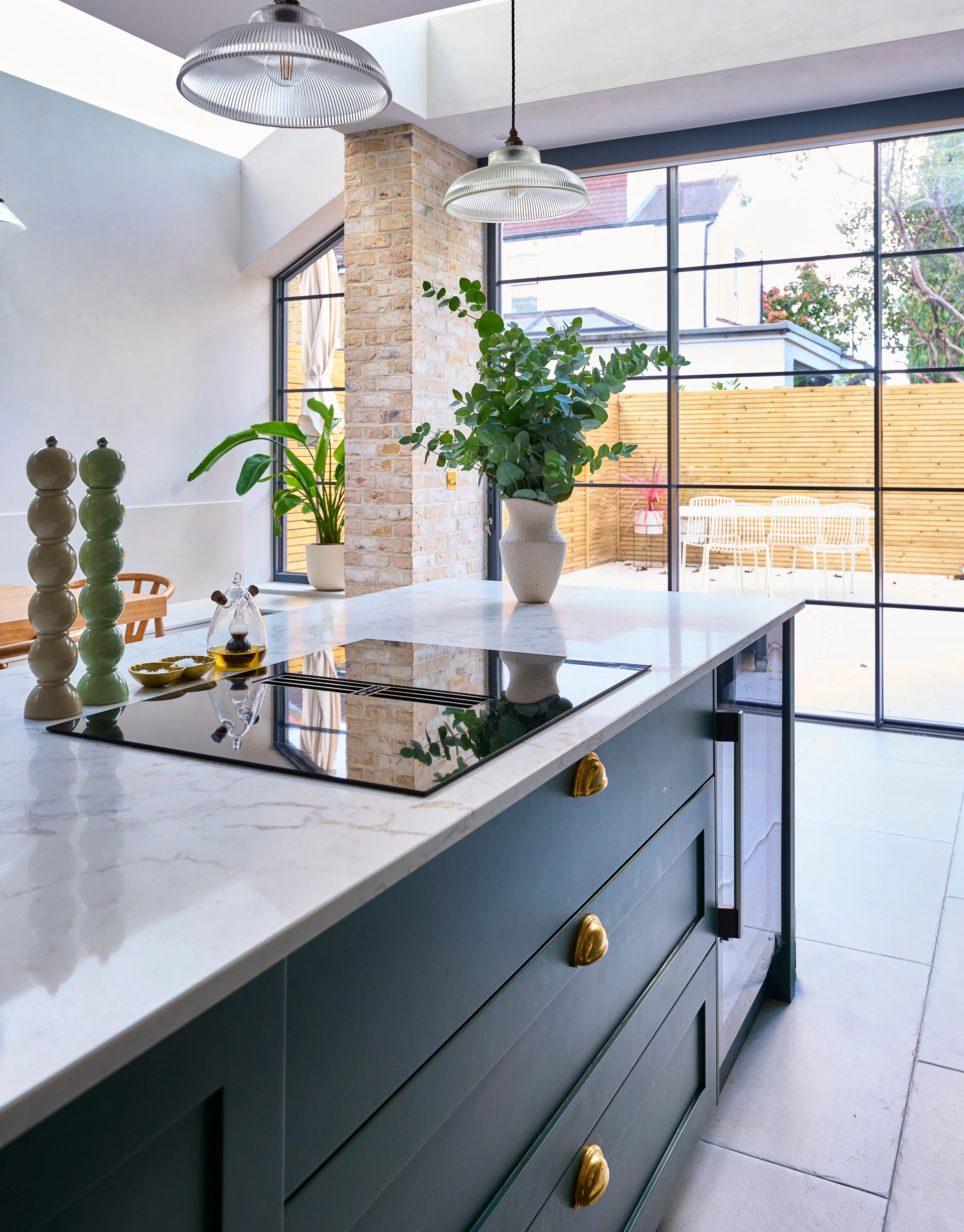 A modern kitchen with a marble countertop, dark cabinet drawers with gold handles, a white vase with green leafy plants, a small glass oil bottle, and a decorative ceramic piece. Large windows reveal an outdoor patio with wooden fencing and outdoor s