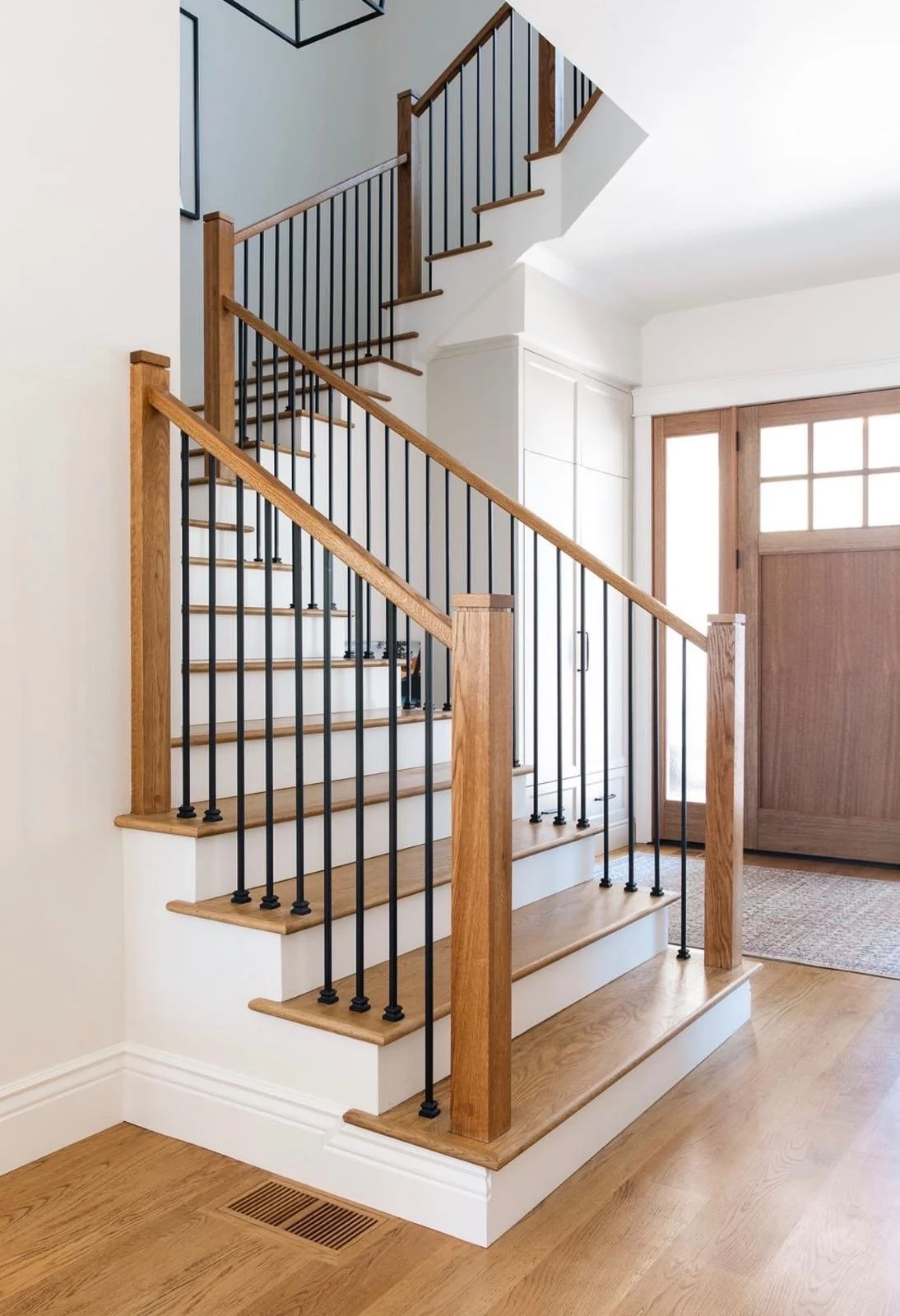 Interior view of a staircase with wooden handrails and black metal balusters, leading to an upper floor. The area has light-colored walls, hardwood flooring, and a large wooden door with glass panels next to the stairs.