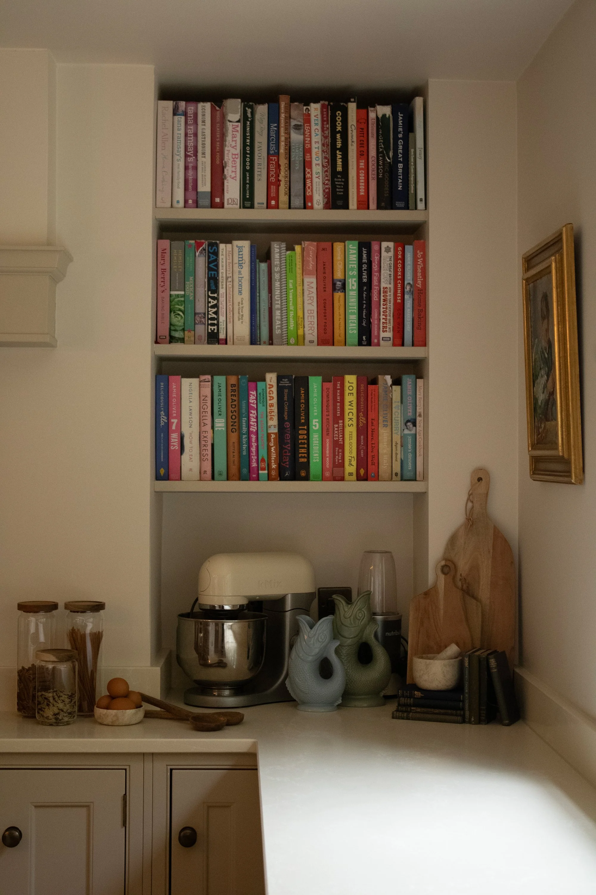 Kitchen shelf with cookbooks, jars with cereal, eggs, kitchen appliances, decorative pottery, and a framed painting on the wall.