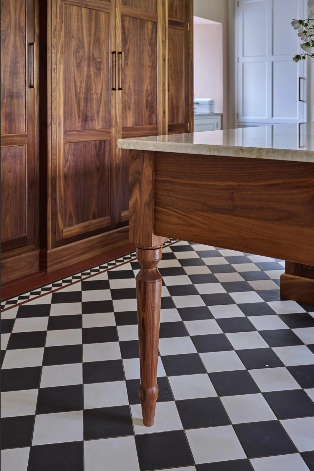 Close-up of a wooden table leg with a marble countertop in a kitchen with wooden cabinets and black-and-white checkered tile flooring.