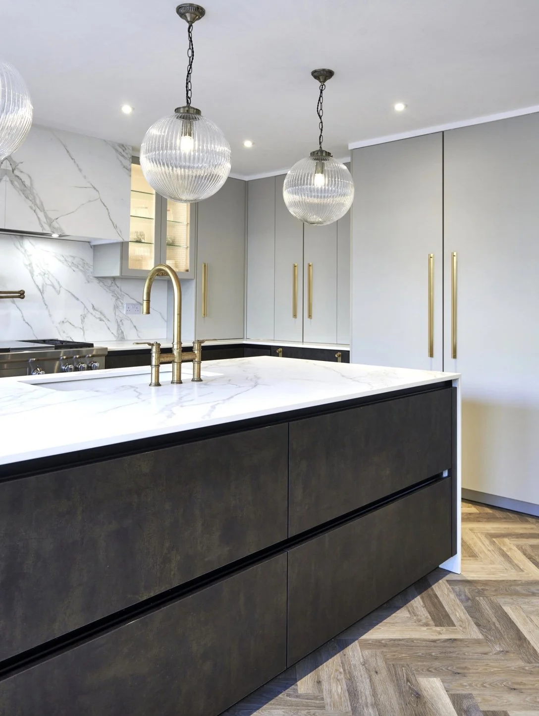 Modern kitchen with white marble countertops, black lower cabinets, light gray upper cabinets with gold handles, hanging pendant lights, and patterned wooden flooring.
