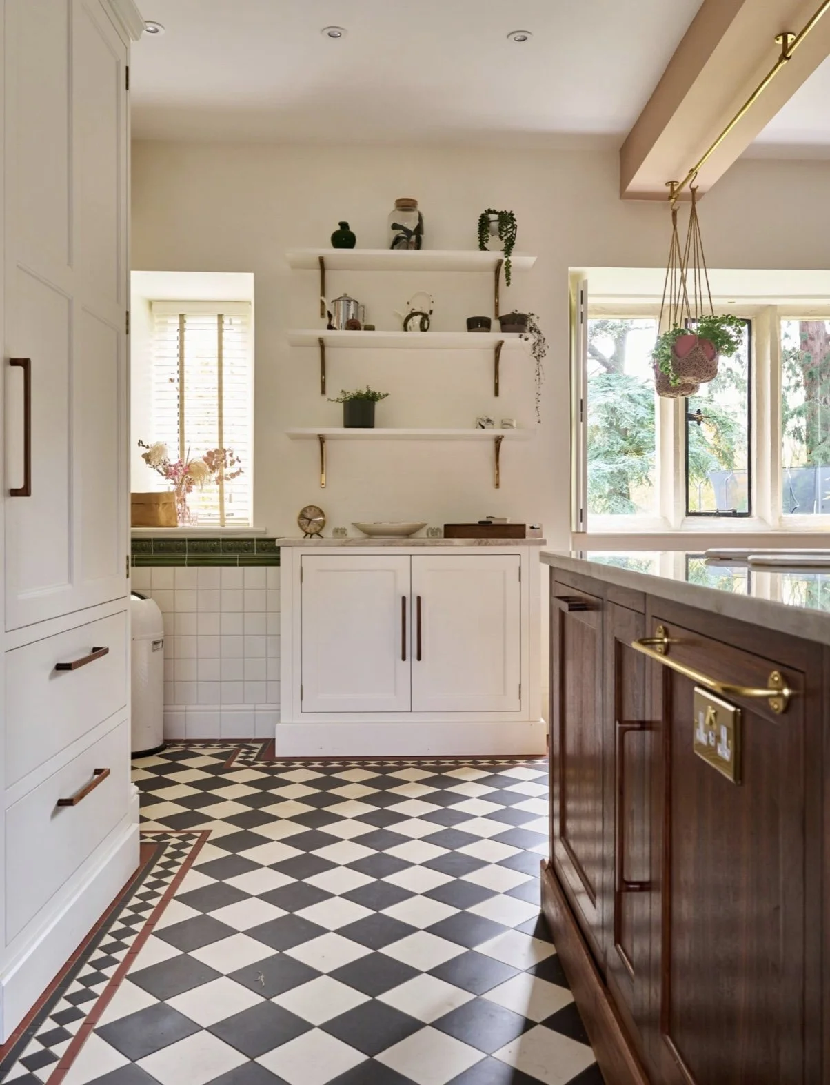 A cozy kitchen with black and white checkered floor tiles, white cabinets, open shelves, and natural light from the windows. Green plants and decorative items are on the shelves and windowsills.