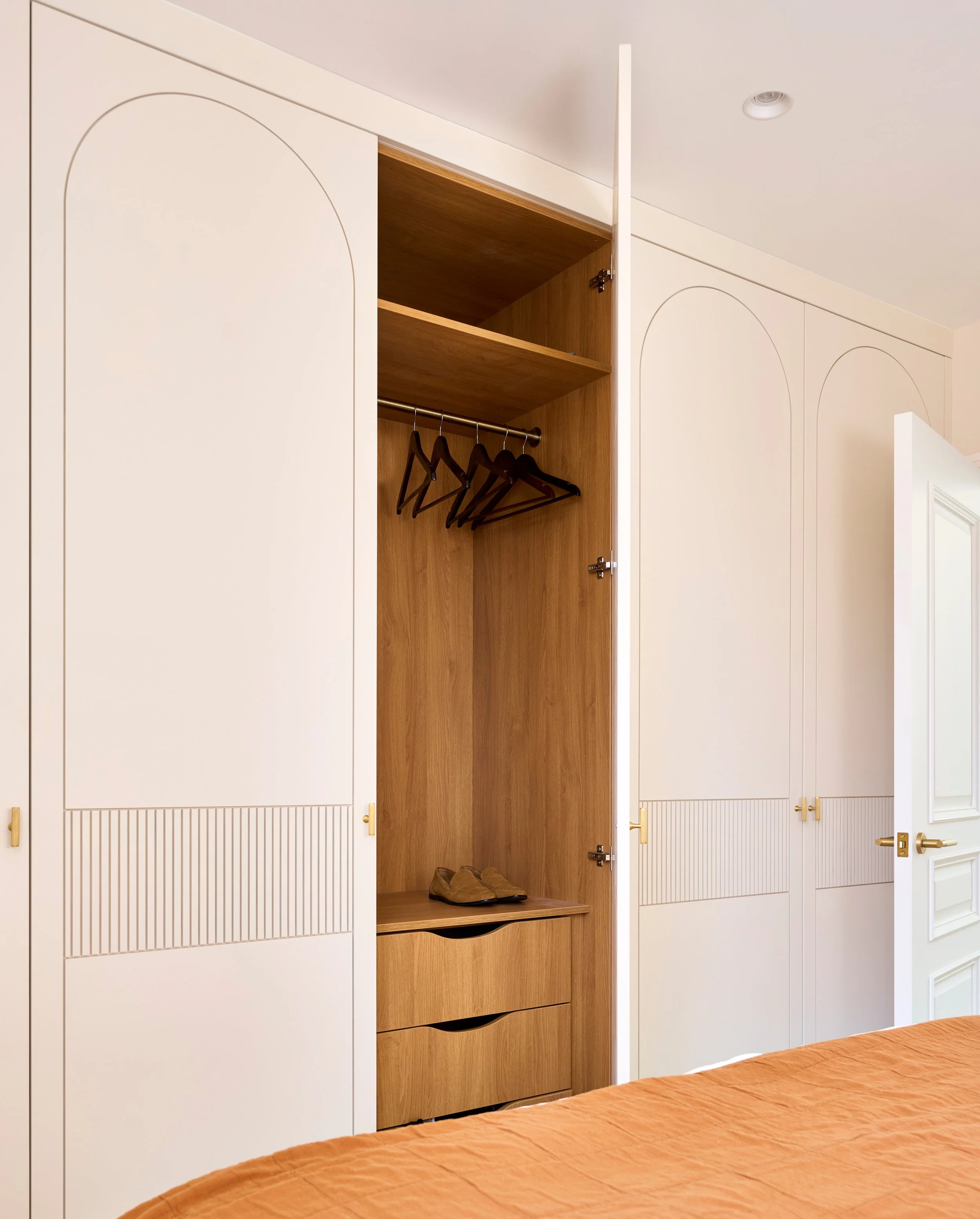 A closet with white doors and a wooden interior, showing shelves and hanging space with black hangers. Shoes are at the bottom. Part of a bed with a brown bedspread is in the foreground.
