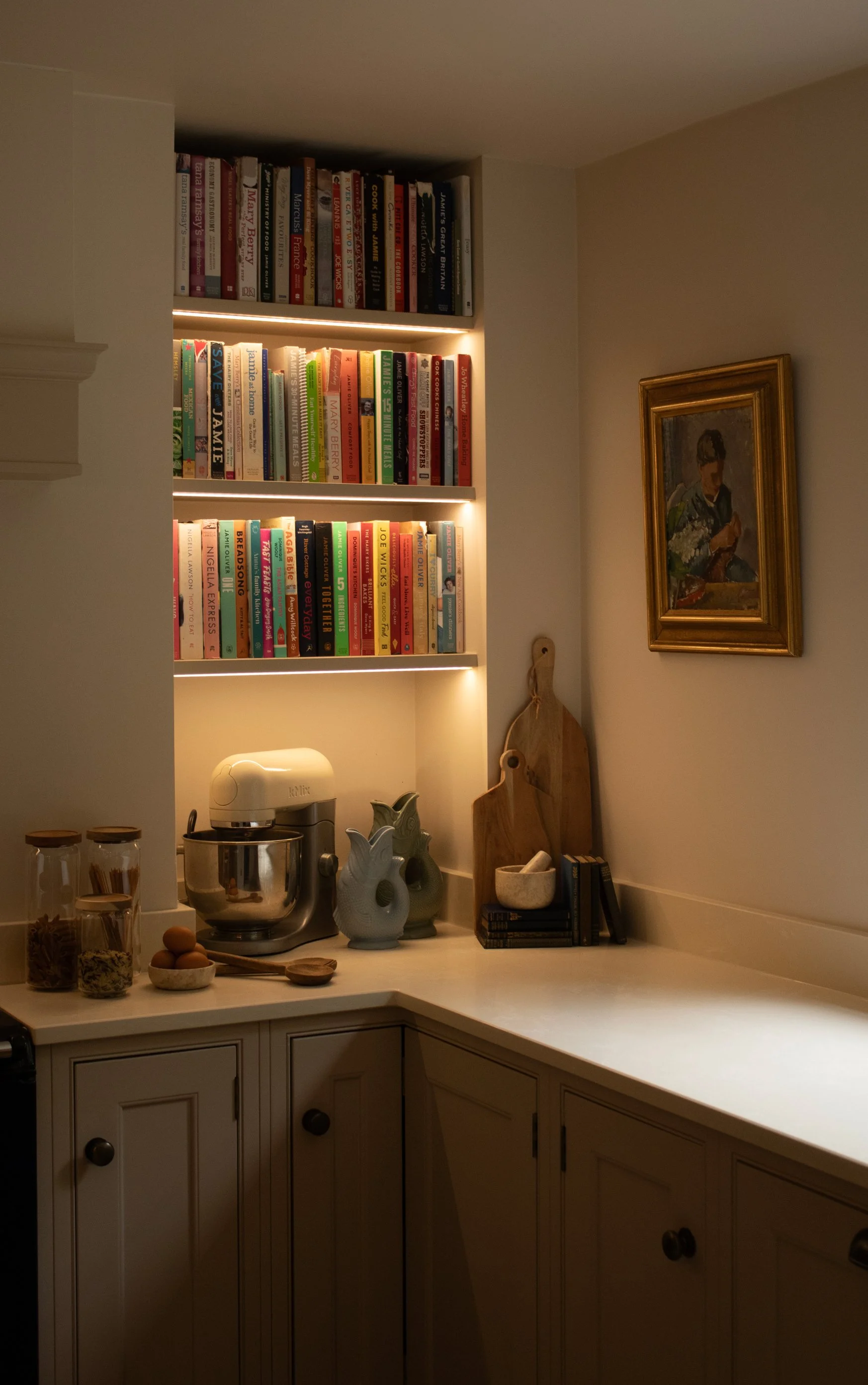 A kitchen corner with a white countertop, a mixer, glass jars, decorative vases, and a baking tray. Above, three shelves hold colorful cookbooks, and a framed painting hangs on the wall.