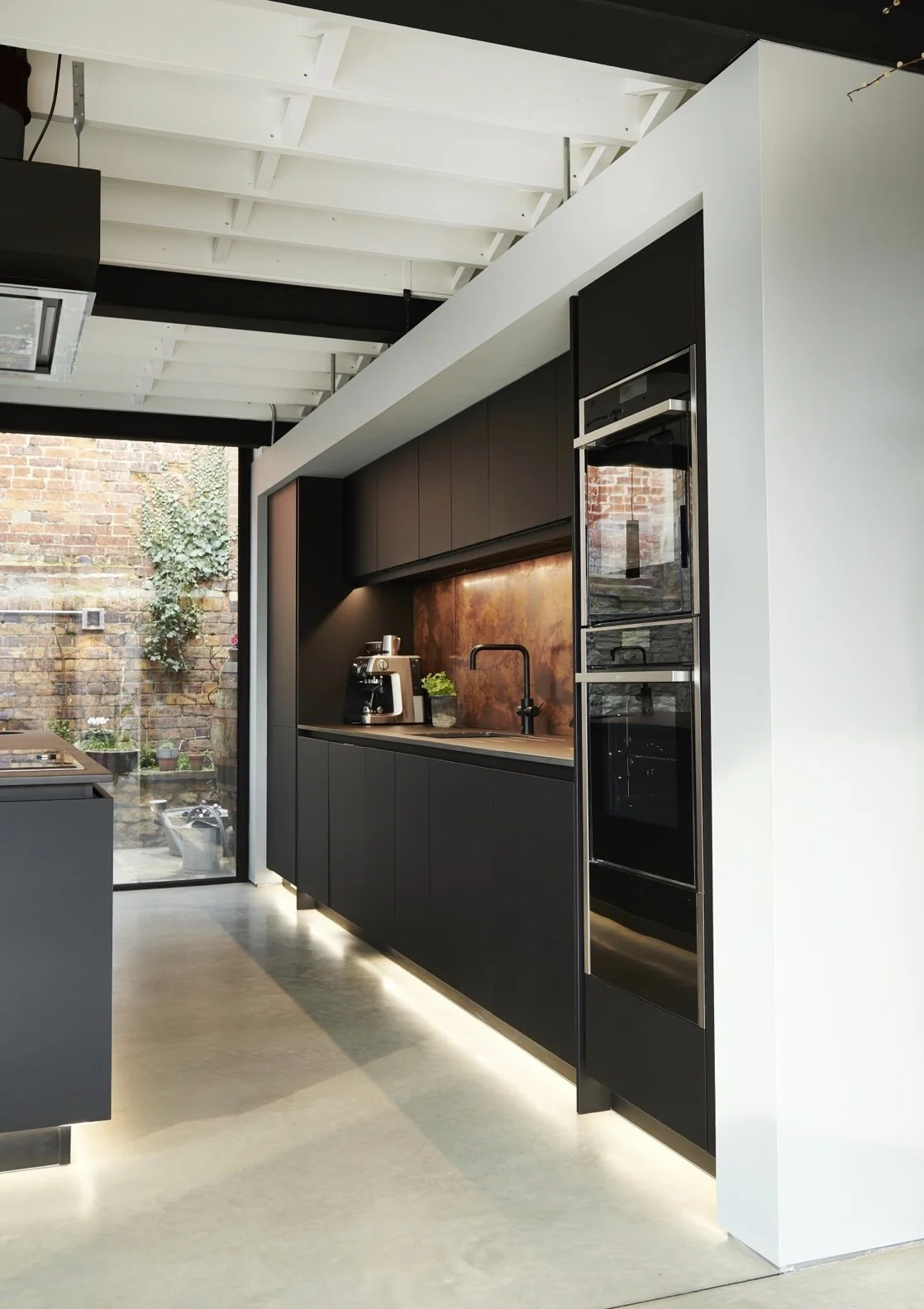Modern black kitchen with built-in oven, coffee maker, black cabinetry, copper backsplash, and concrete flooring, with a view of an outdoor brick wall.