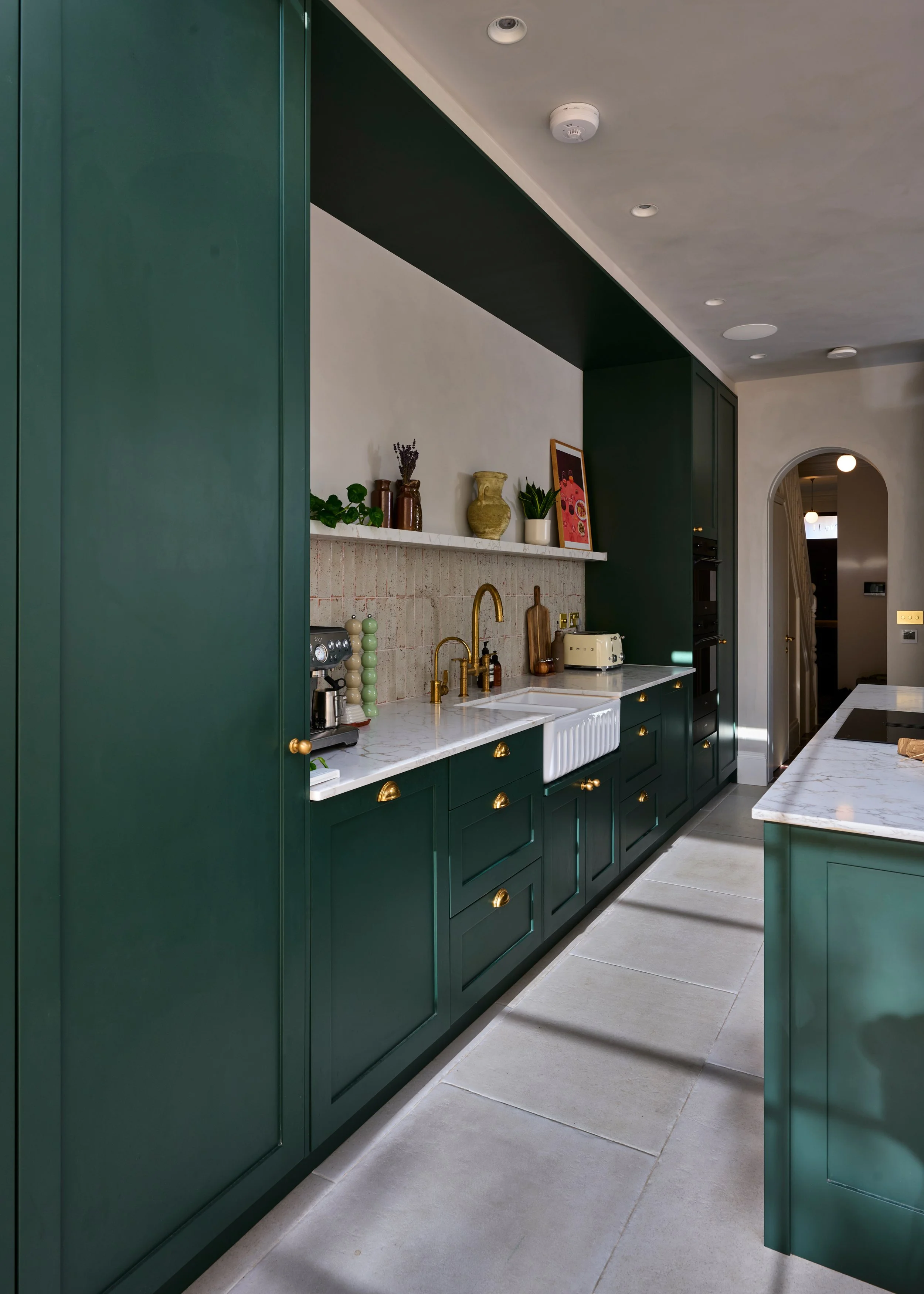Modern kitchen with green cabinets, gold hardware, white marble countertops, and a white farmhouse sink. Decor includes vases, plants, and framed art on a shelf, with a doorway leading to another room.