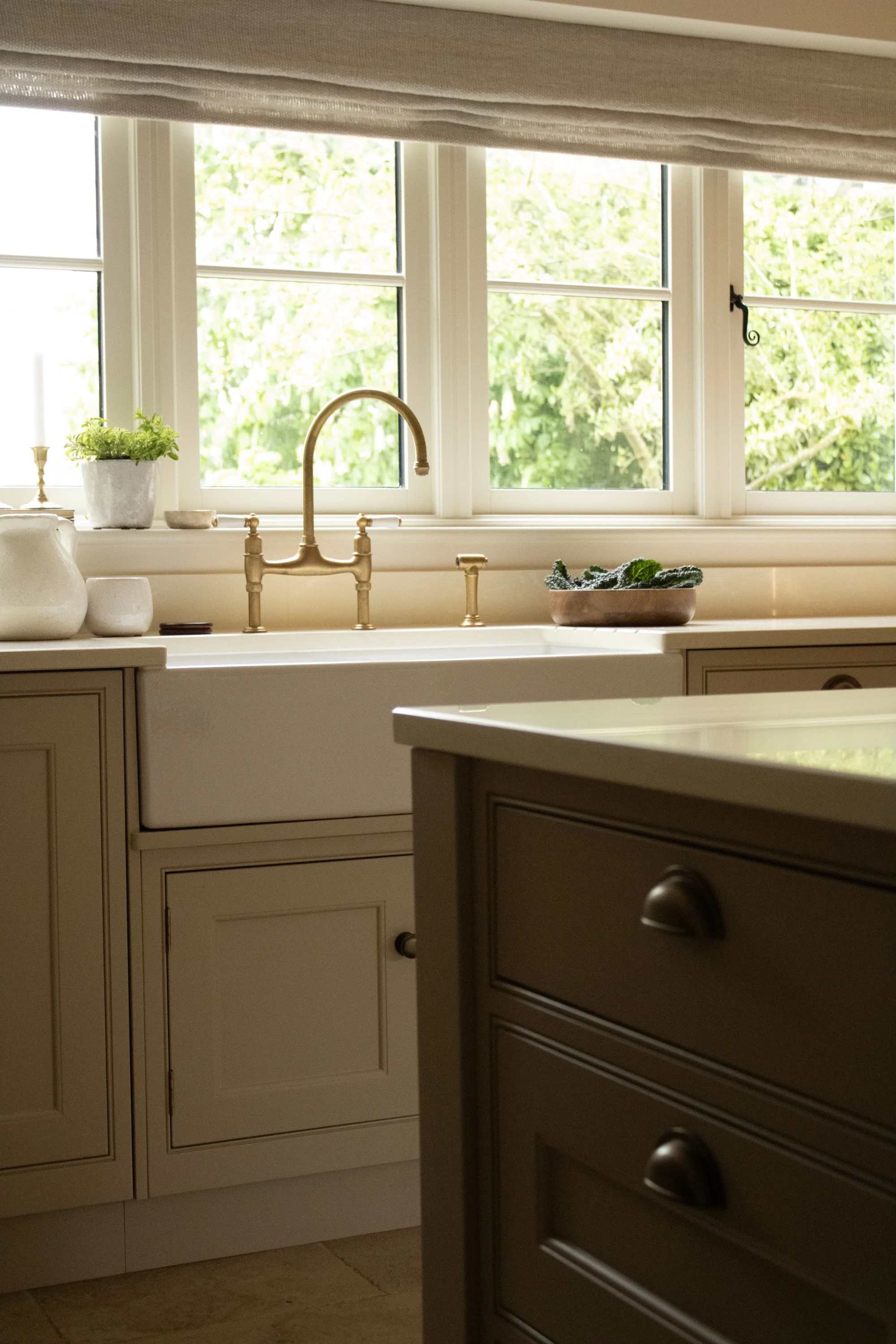 A kitchen with a window above the sink, showing greenery outside. The window has a roller shade partly drawn. On the countertop, there is a potted plant, a bowl, and some small containers. The cabinetry is light-colored with classic paneling.