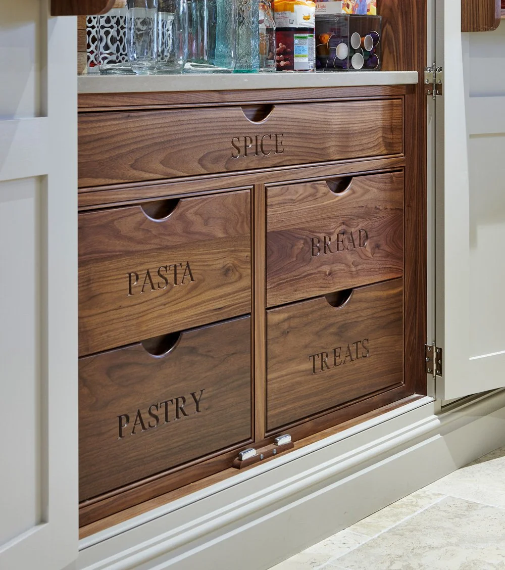 Wooden drawers labeled 'SPICE,' 'PASTA,' 'PASTRY,' 'BREAD,' and 'TREATS' inside a kitchen cabinet, with a cream-colored countertop above and various glass containers and coffee capsules on top.