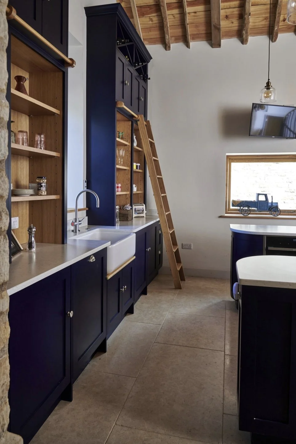 A rustic kitchen with navy blue cabinets, open wooden shelves, a white farmhouse sink, and a wooden ladder leaning against tall cabinets. Natural light enters through a window decorated with a toy truck, with exposed wooden ceiling beams and a hangin