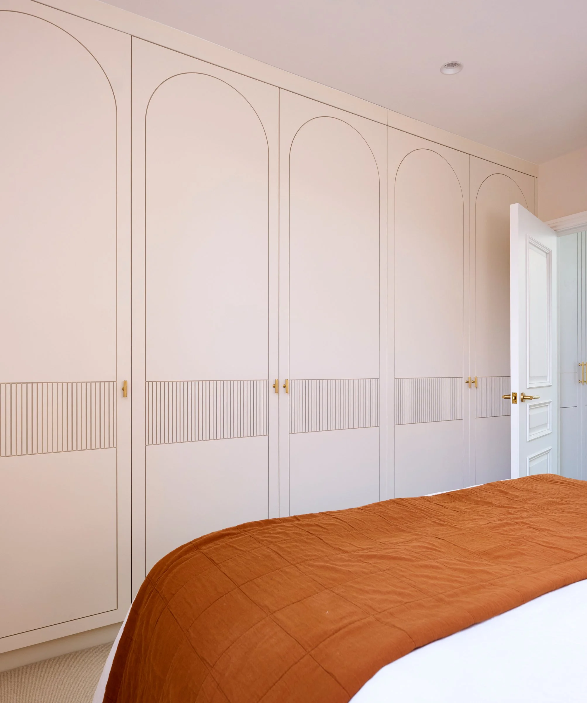 A bedroom with a white wardrobe with gold handles, a beige carpet, and part of a bed with a brown quilted bedspread visible.
