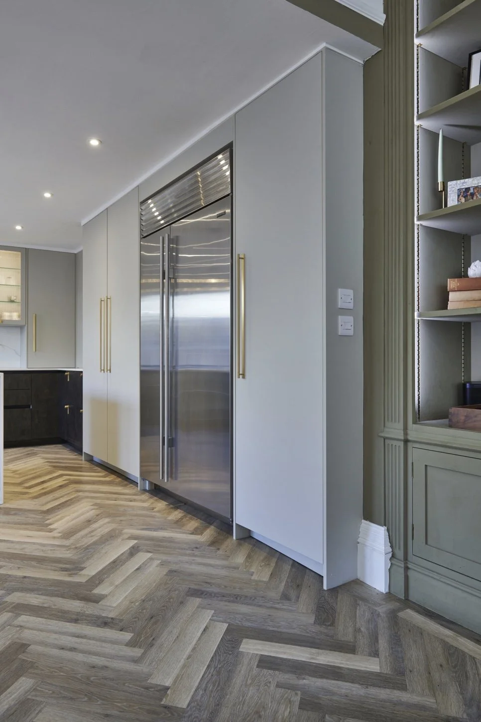 Modern kitchen with built-in stainless steel refrigerator, white cabinetry with gold handles, light green shelving unit, and herringbone patterned hardwood floor.