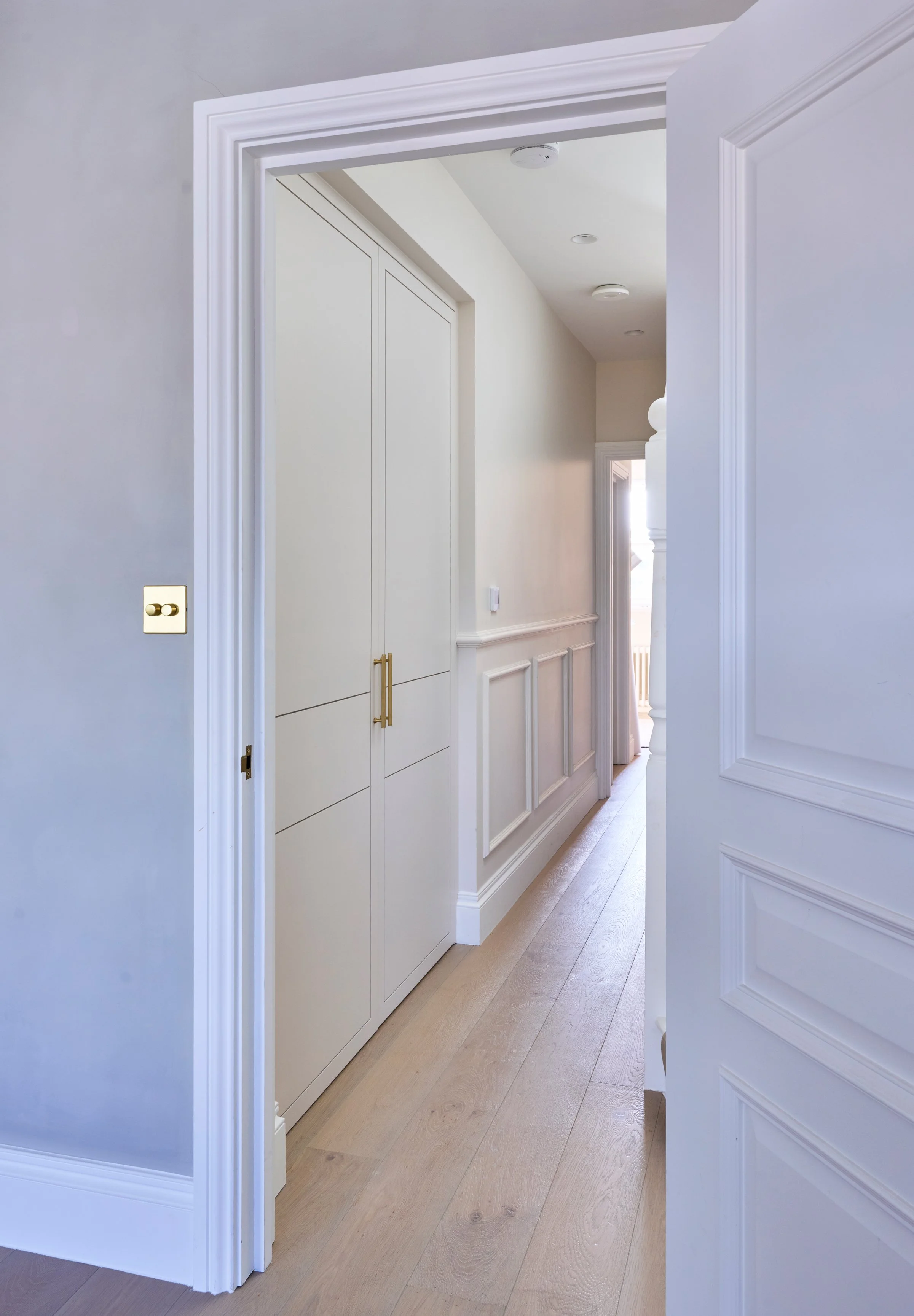 View through a doorway into a hallway with white walls, light wood flooring, and a built-in closet with gold handles.