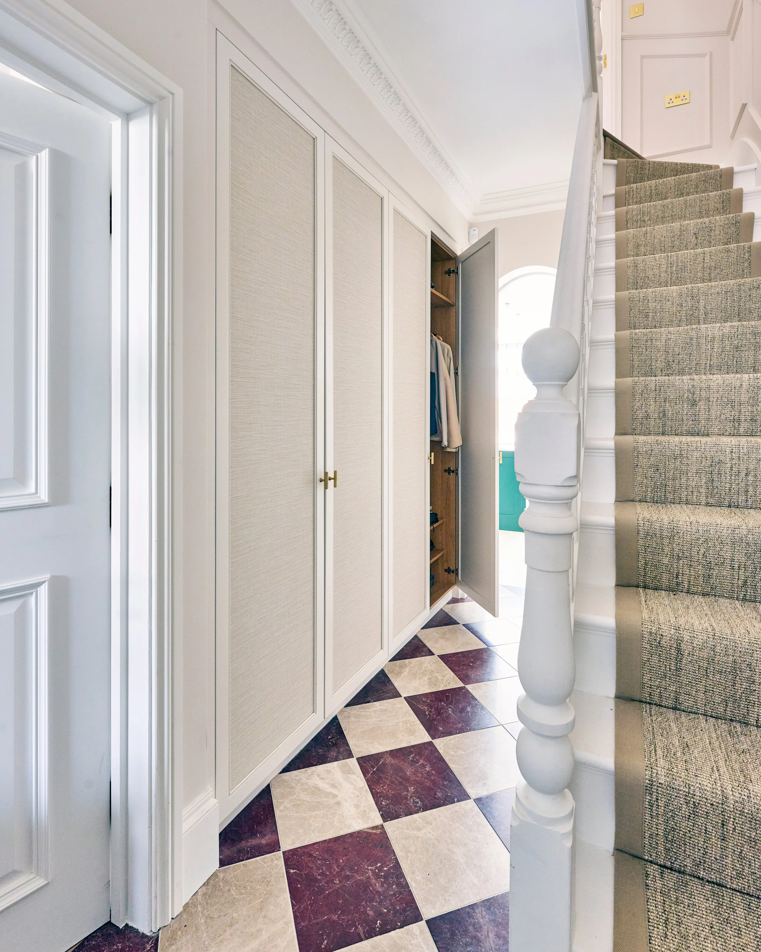 Interior of a house entryway with a staircase, beige and dark marble patterned flooring, built-in cabinetry, and a door opening to another room.