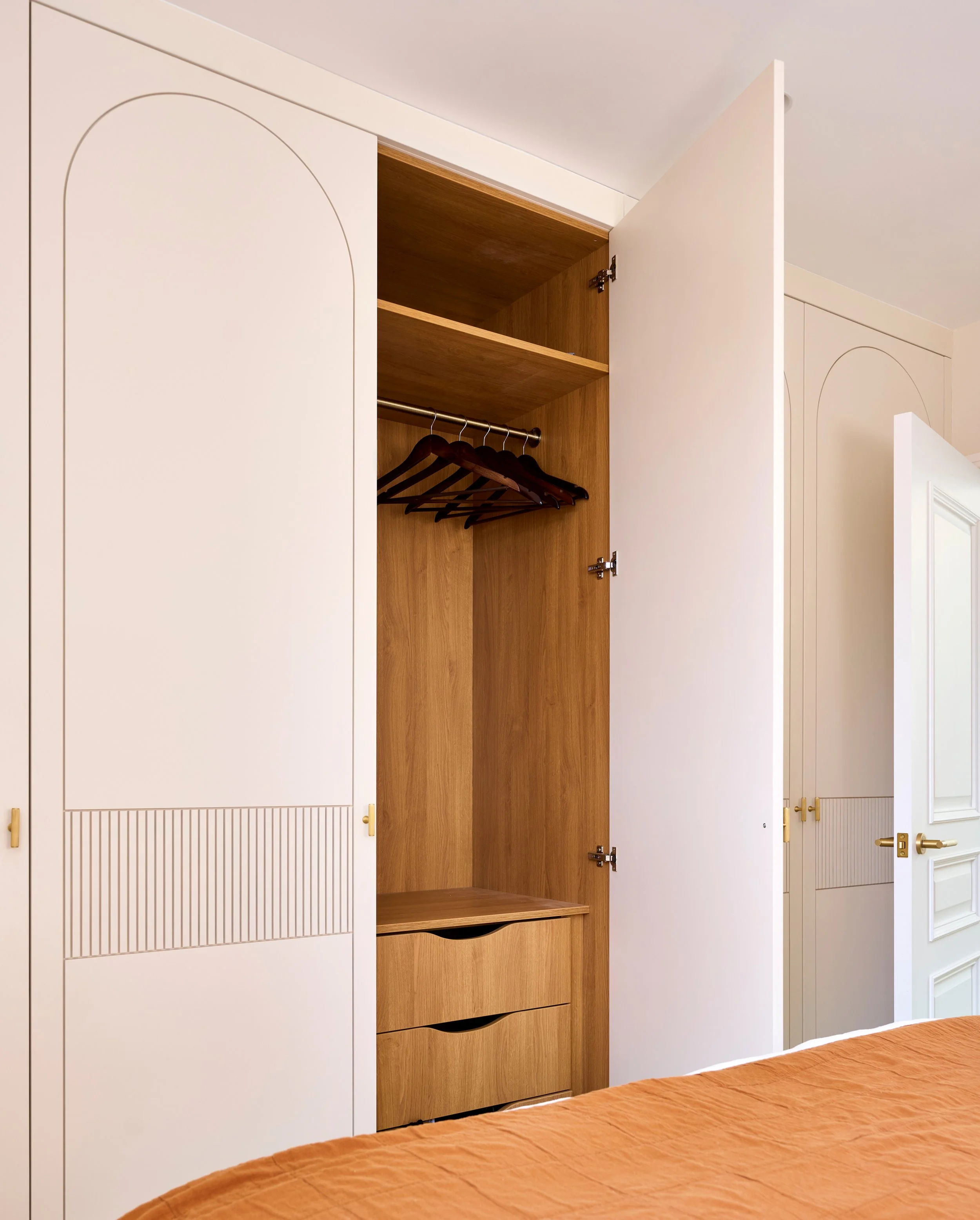 Open wardrobe with wooden interior, hanging rods, and drawers, in a room with white paneled walls and a bed with an orange bedspread.