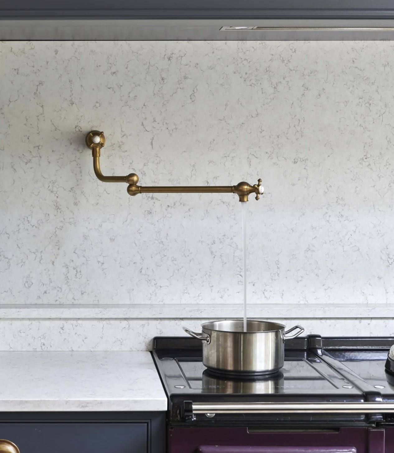 A kitchen with a brass pot filler faucet mounted on the wall above a stove, with water flowing into a stainless steel pot.