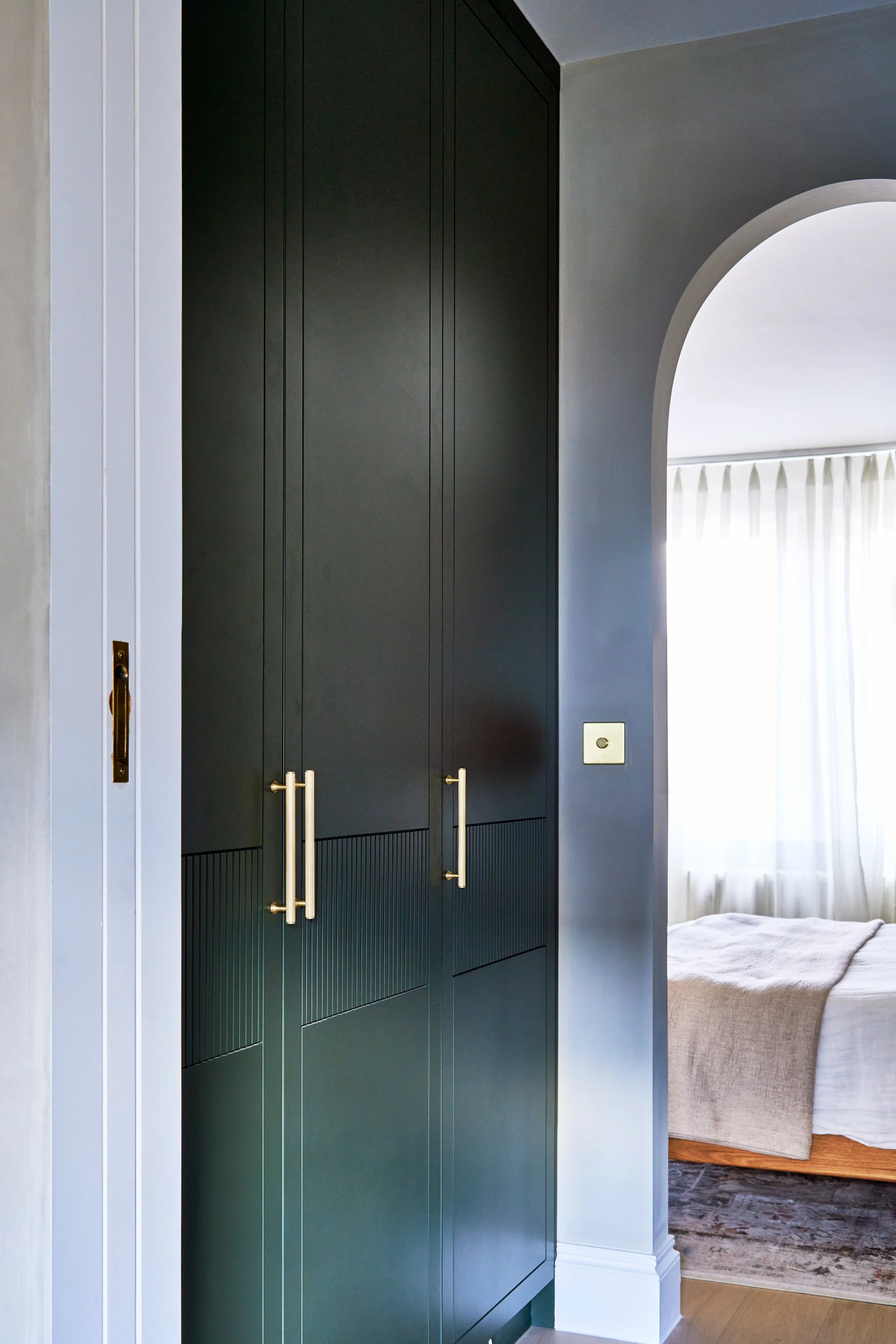 A hallway with dark green cabinets featuring gold handles on the left, part of a white door frame and wall on the far left, and an arched doorway leading into a bedroom with a window covered by light curtains and a bed with a beige cover on the right
