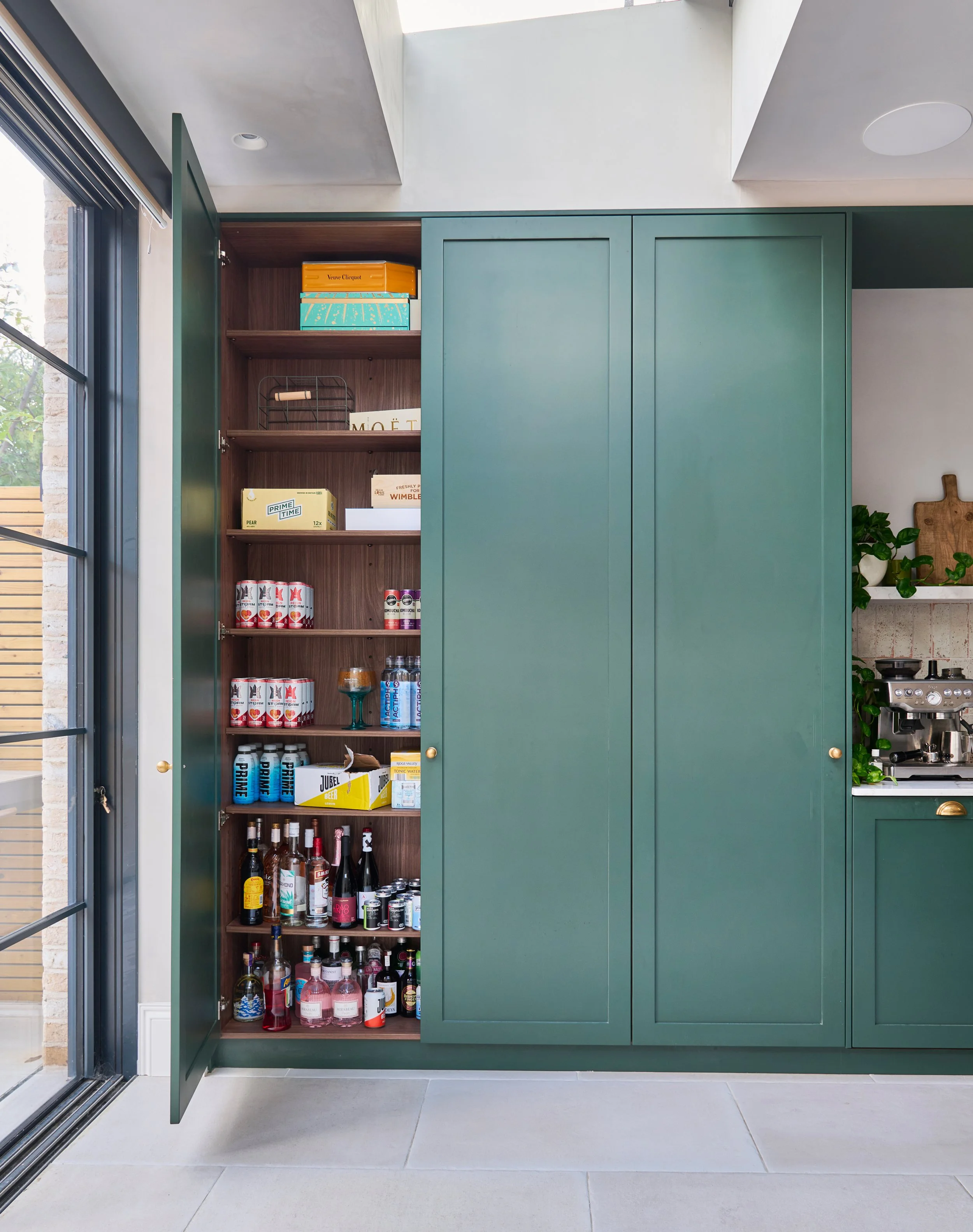 A kitchen cabinet with one door open showing bottles, cans, and boxes of various beverages and food items inside. The cabinet is painted green, and there is a window with black frames on the left side of the image, allowing natural light into the roo