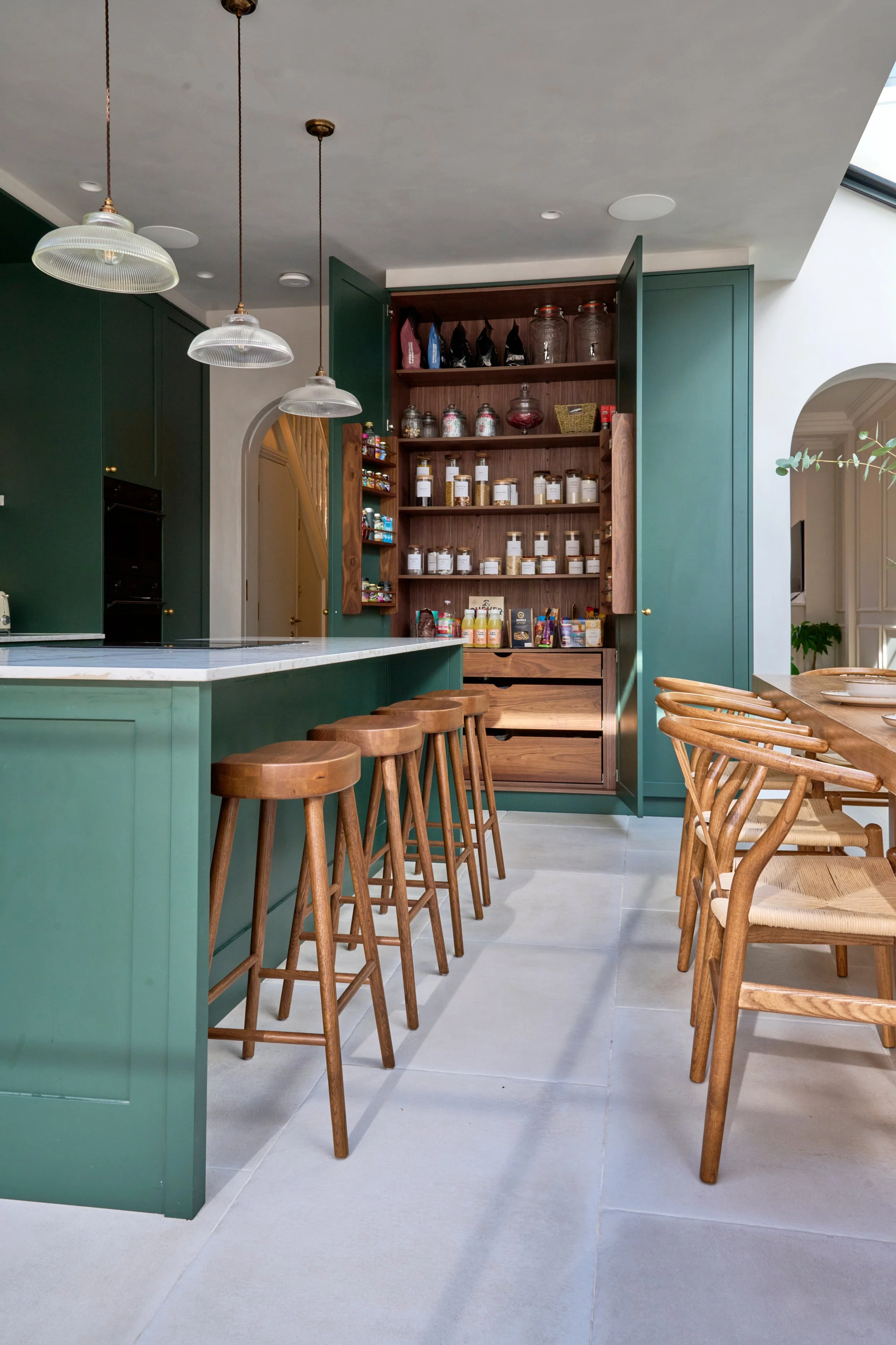 View of a modern kitchen with green cabinets, a marble countertop, wooden stools, and open shelving with jars and pantry items. Bright natural light streams in from a skylight or window on the right.