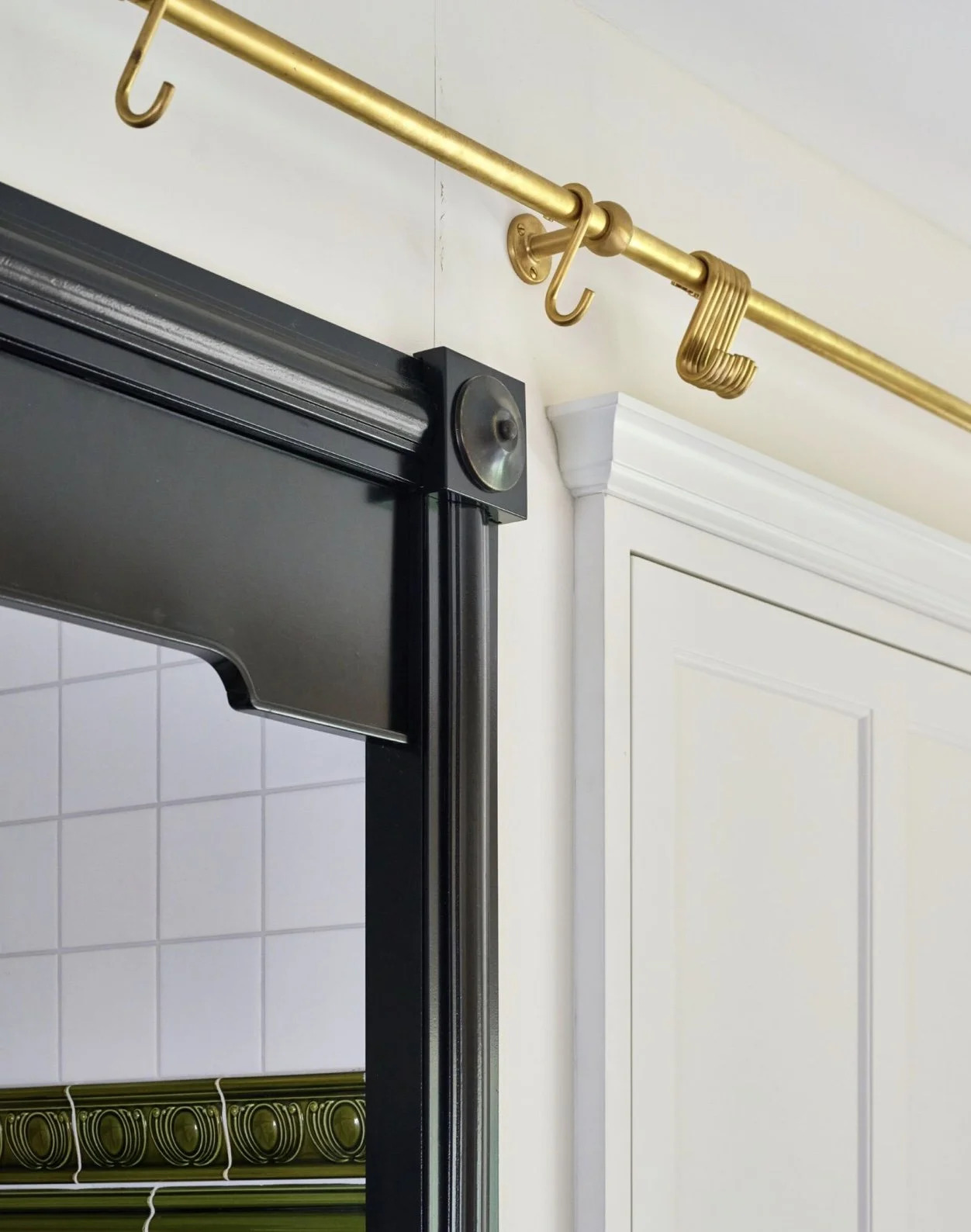 Close-up of a black shower door frame with a round roller and a modern gold curtain rod with hooks, white wall, and white cabinetry.