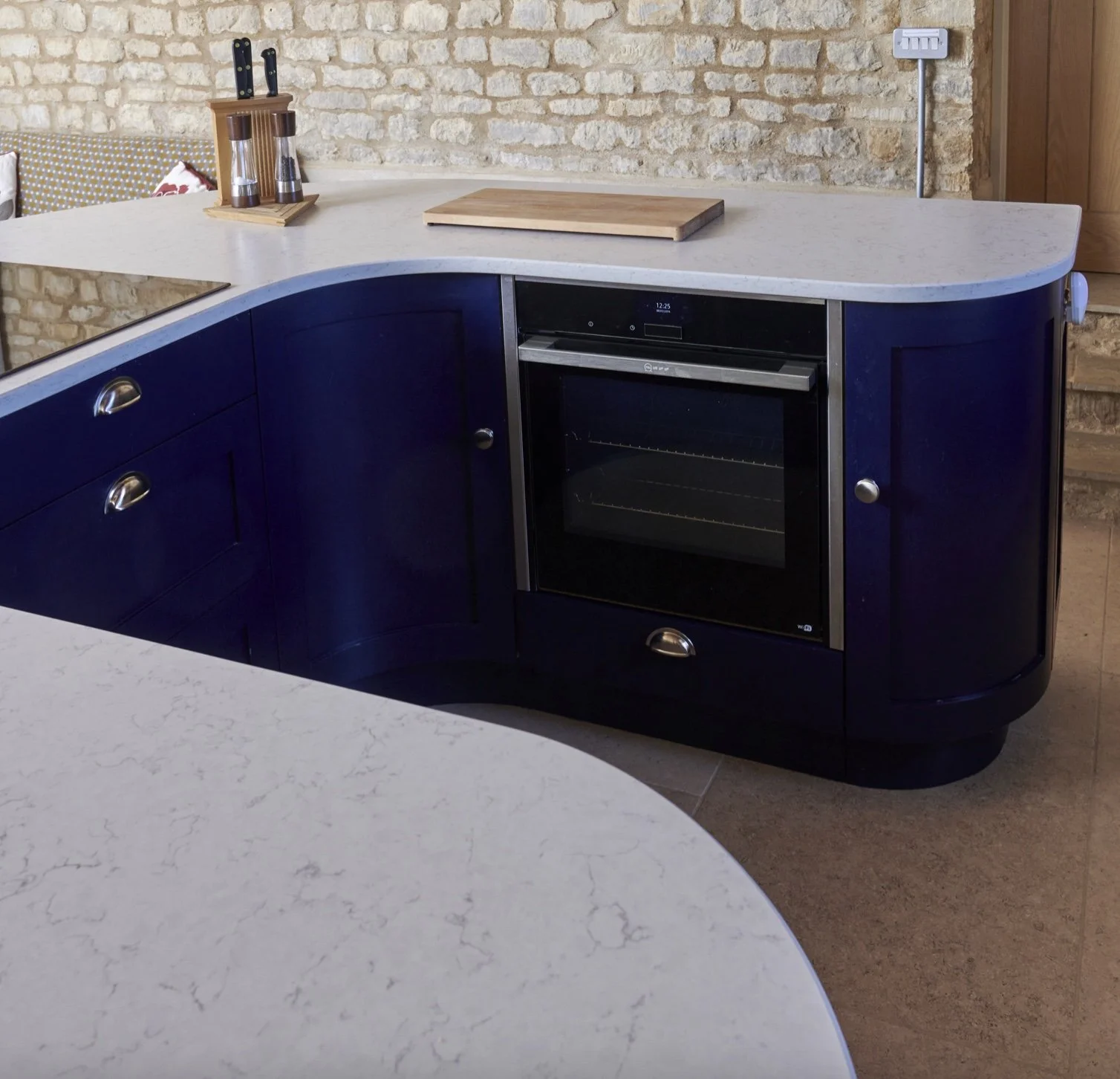 A kitchen with a curved navy blue cabinet, a white countertop, and an oven. There is a wooden cutting board on the counter and a knife block with knives. The background features a brick wall and a wooden panel.