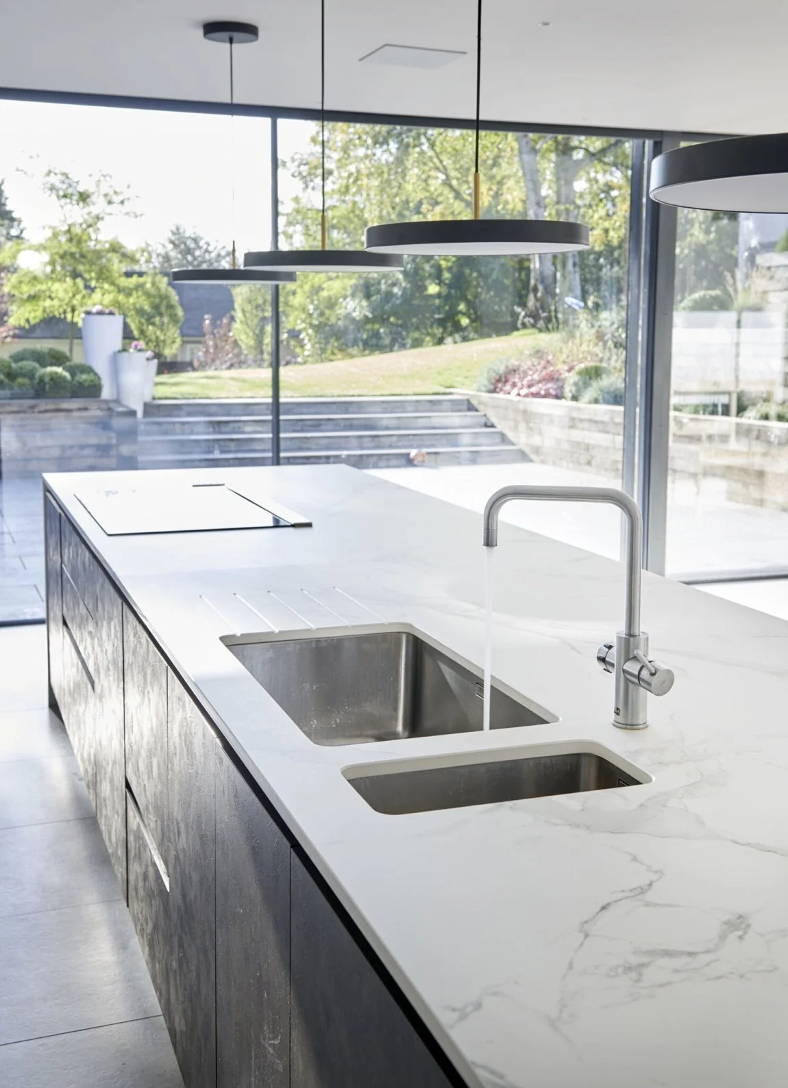 Modern kitchen island with white marble countertop, dual stainless steel sinks, and a sleek faucet, with large windows and a view of an outdoor garden.