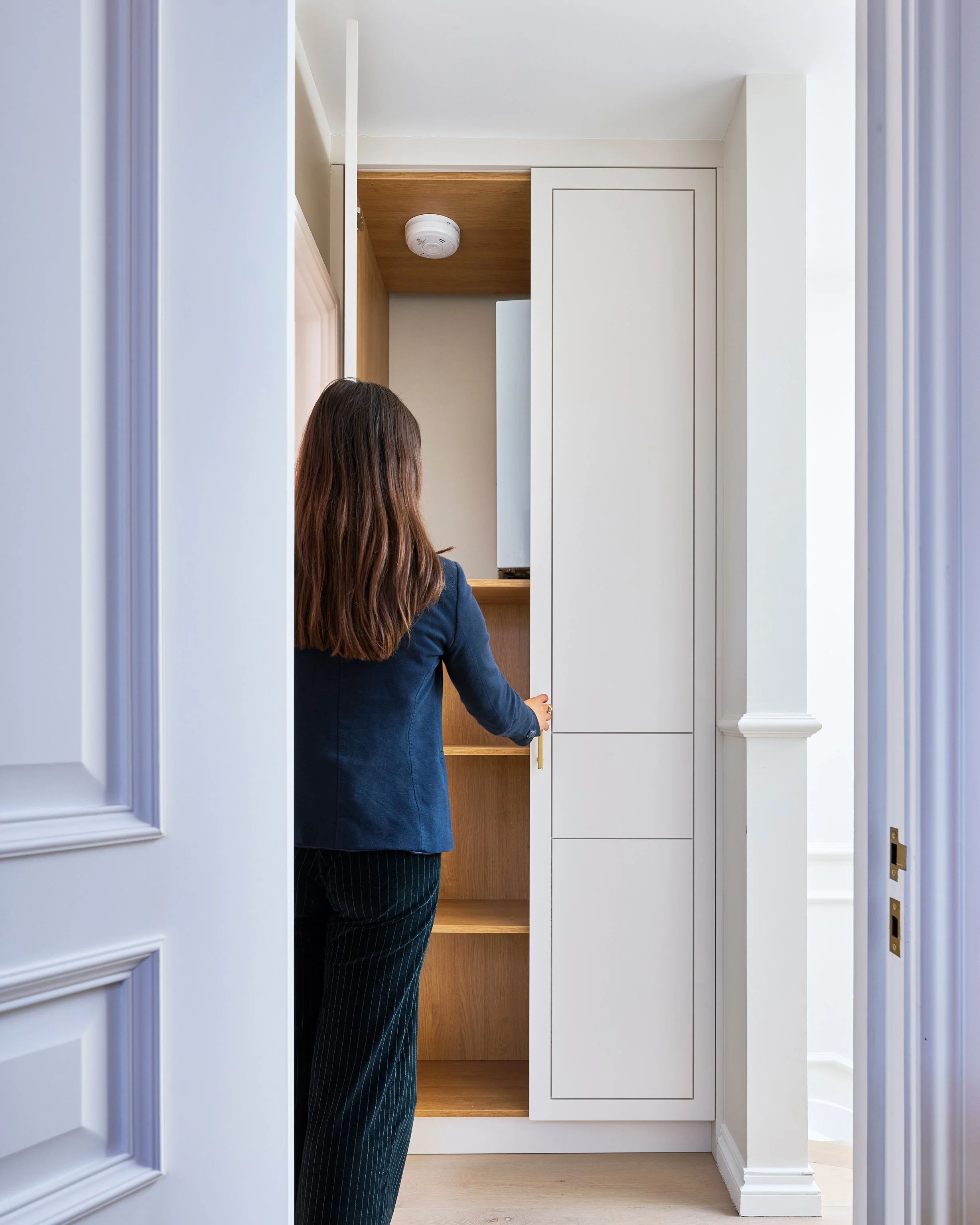 A woman with long brown hair, dressed in a blue blazer and pinstripe pants, opening a white cabinet door in a room with white walls and light wood flooring.