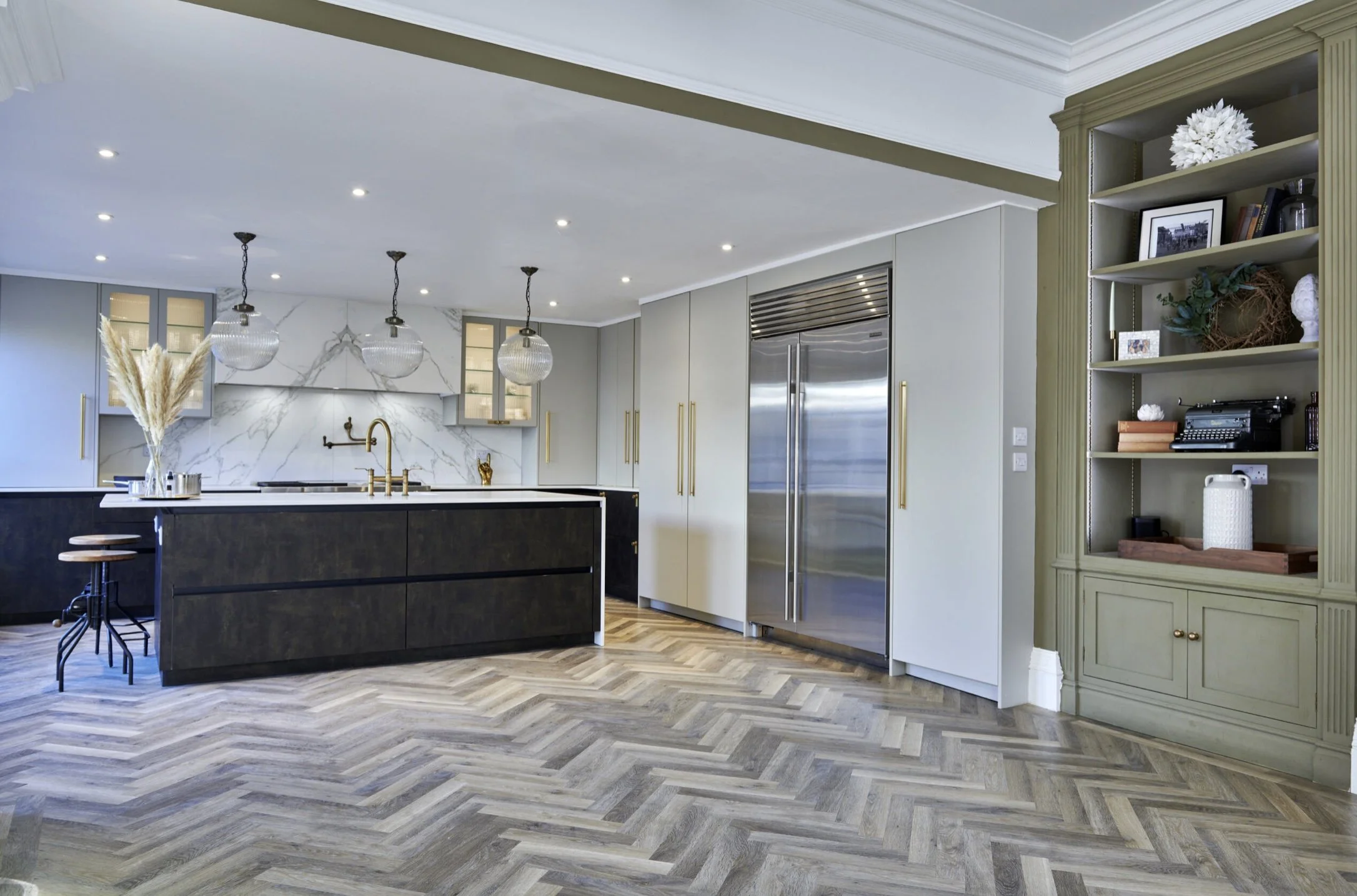 Modern kitchen with black island, white marble backsplash, gold fixtures, gray cabinets, stainless steel refrigerator, and green built-in bookshelf.