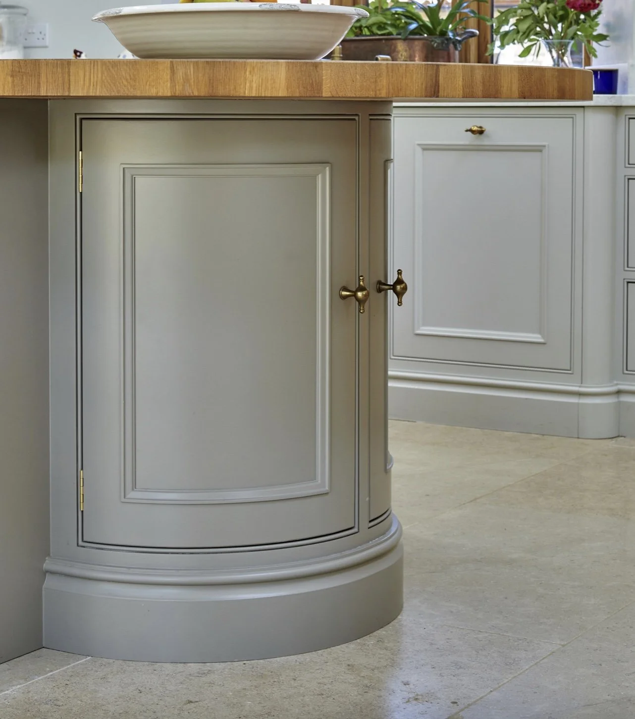 Close-up of a kitchen island with a painted grey cabinet door with brass hardware, a wooden countertop, and some plants in the background.