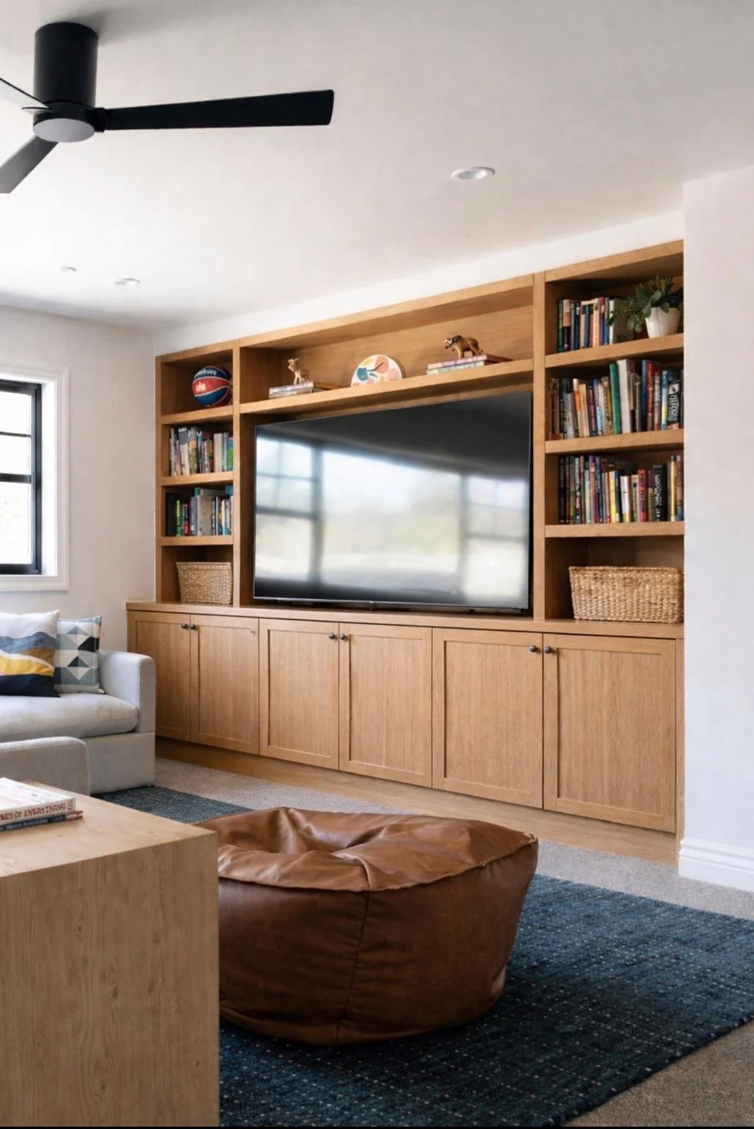 Living room with built-in wooden shelves, a flat-screen TV, a cream sofa with patterned pillows, a brown leather bean bag chair on a dark rug, a black ceiling fan, and a window with black framing.