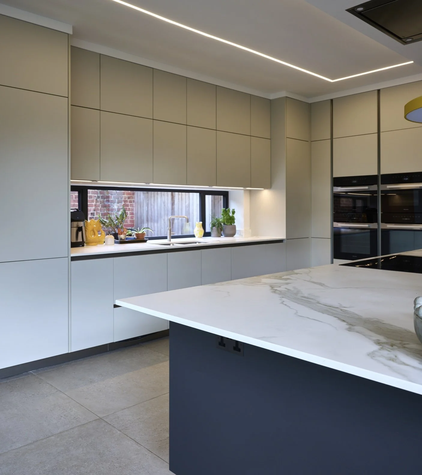Modern kitchen with beige cabinets, marble countertop island, and a window with plants and decorative items.