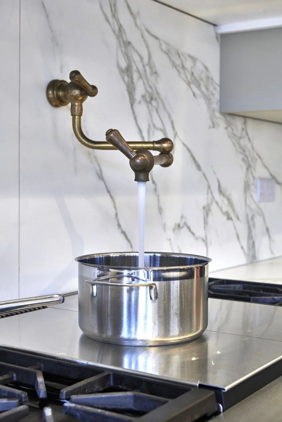 A brass kitchen faucet with water running into a stainless steel pot on a stove, with a marble backsplash in the background.