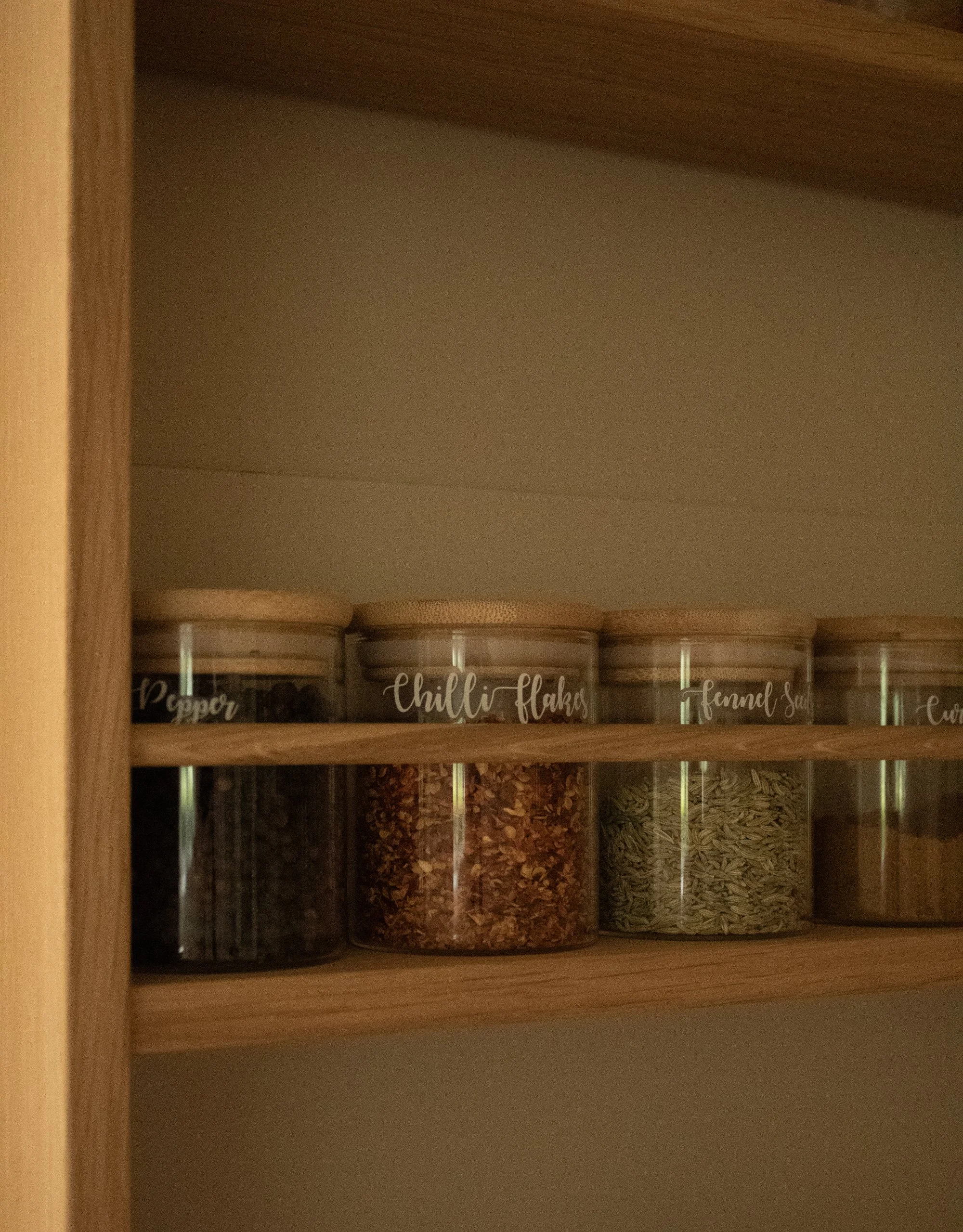 Shelf with clear jars labeled with different spices and seasonings, including chili flakes, fennel seeds, and pepper.