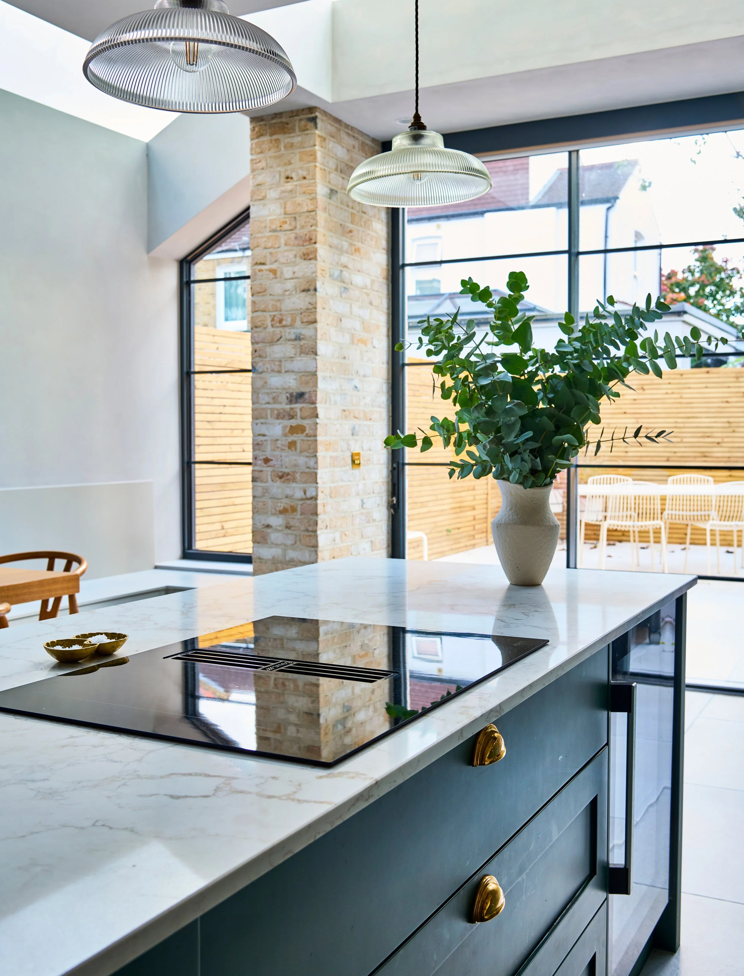 Modern kitchen with white marble countertop, a built-in induction stove, and dark blue cabinets with gold handles. A large vase with green leafy plant sits on the countertop, with a brick wall, large windows, and outdoor wooden fencing visible in the