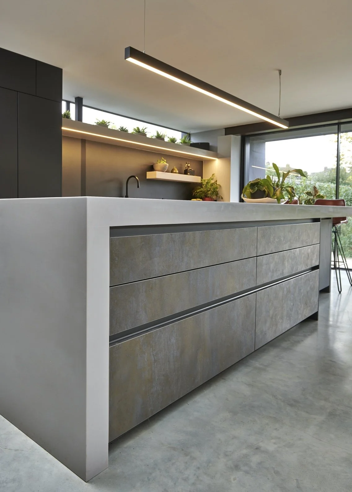 Modern kitchen with a white island countertop, gray drawers, and black cabinetry. A black faucet, potted plants, and decorative items are on the countertop. Large windows and black and white shelves with plants and decorations are in the background.