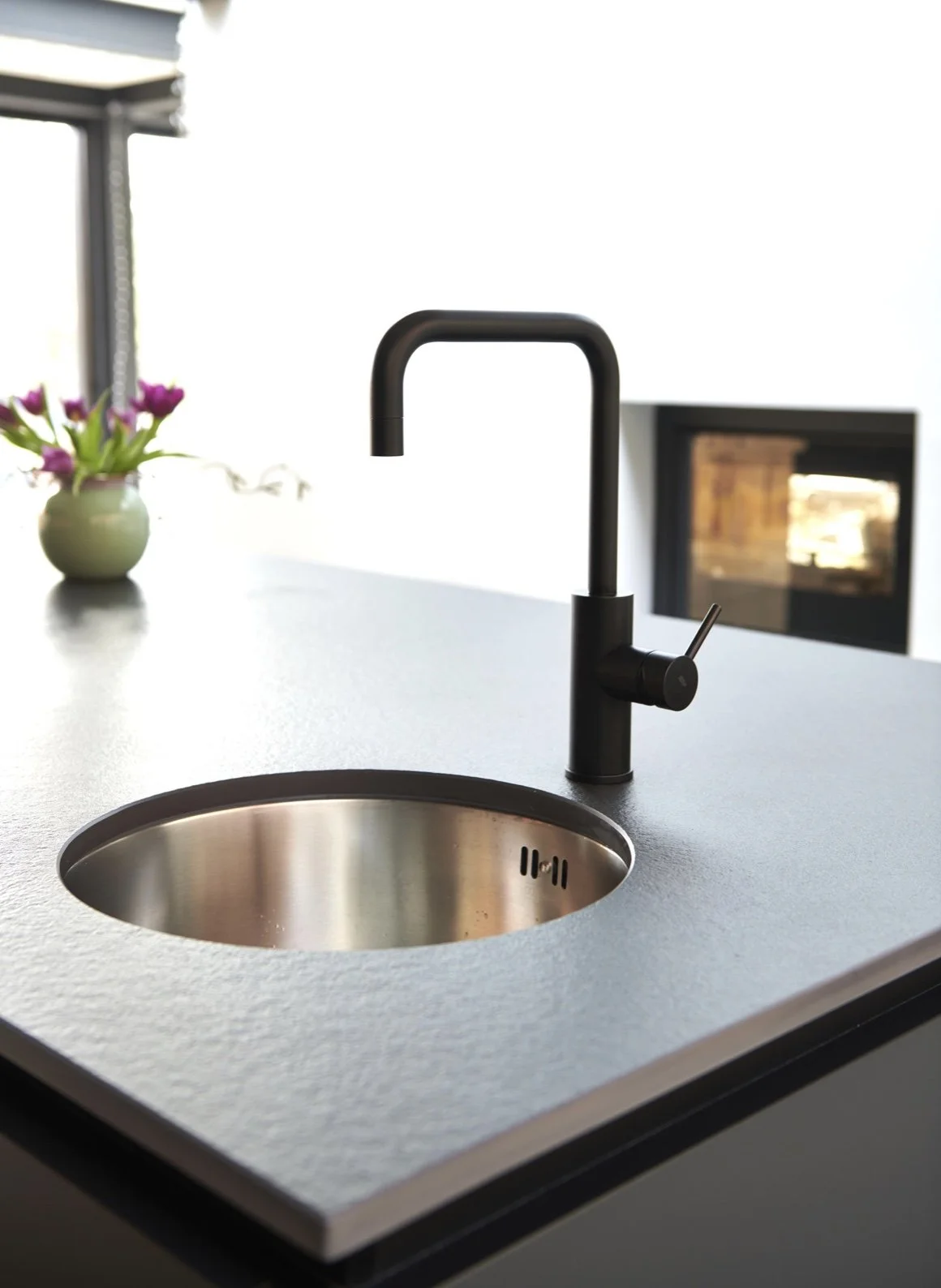 Modern black kitchen faucet with a single handle mounted on a light-colored countertop, with a small flower arrangement in a white vase and a fireplace in the background.