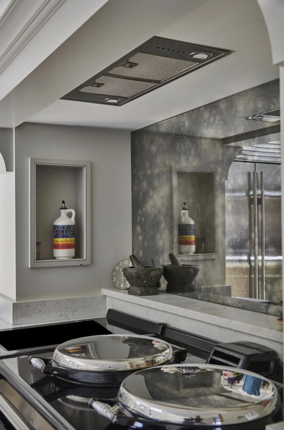 A modern kitchen with a metallic stovetop, decorative bowls, wall-mounted shelf with a colorful vase, and a mirror reflecting part of the kitchen interior.