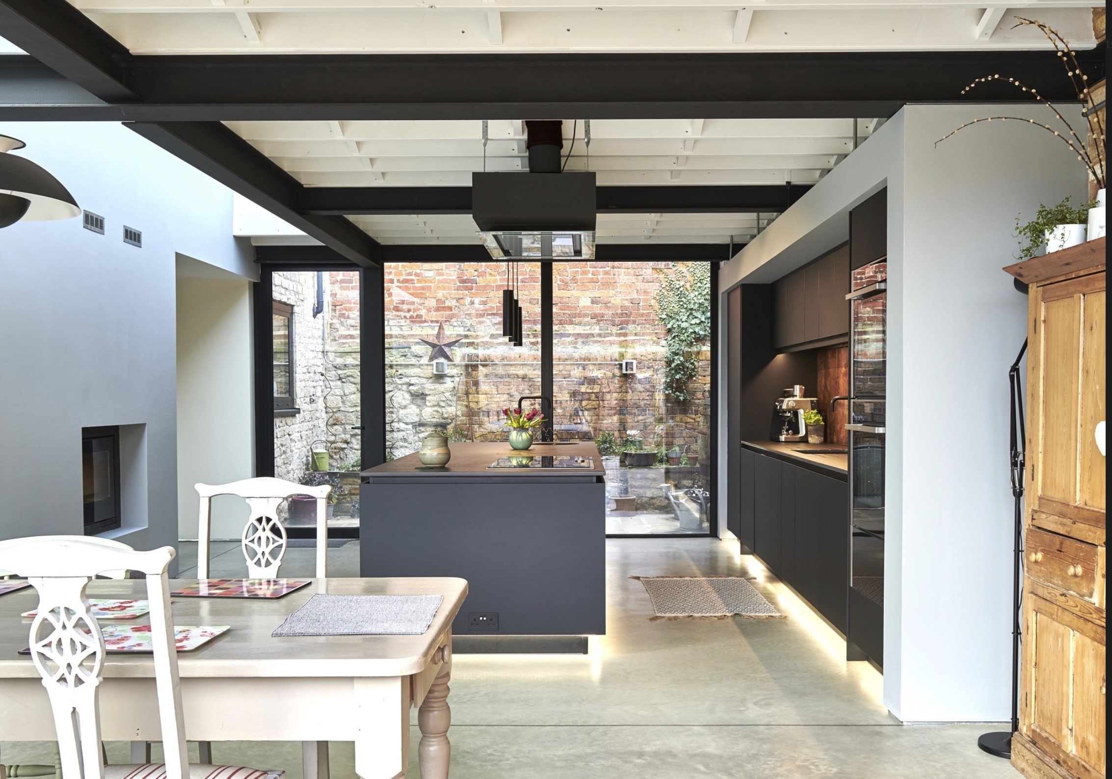 Modern kitchen with black cabinetry, a kitchen island with a sink and flowers, a glass wall overlooking a brick outdoor area, a wooden dining table with white chairs, and a wooden cabinet on the right.