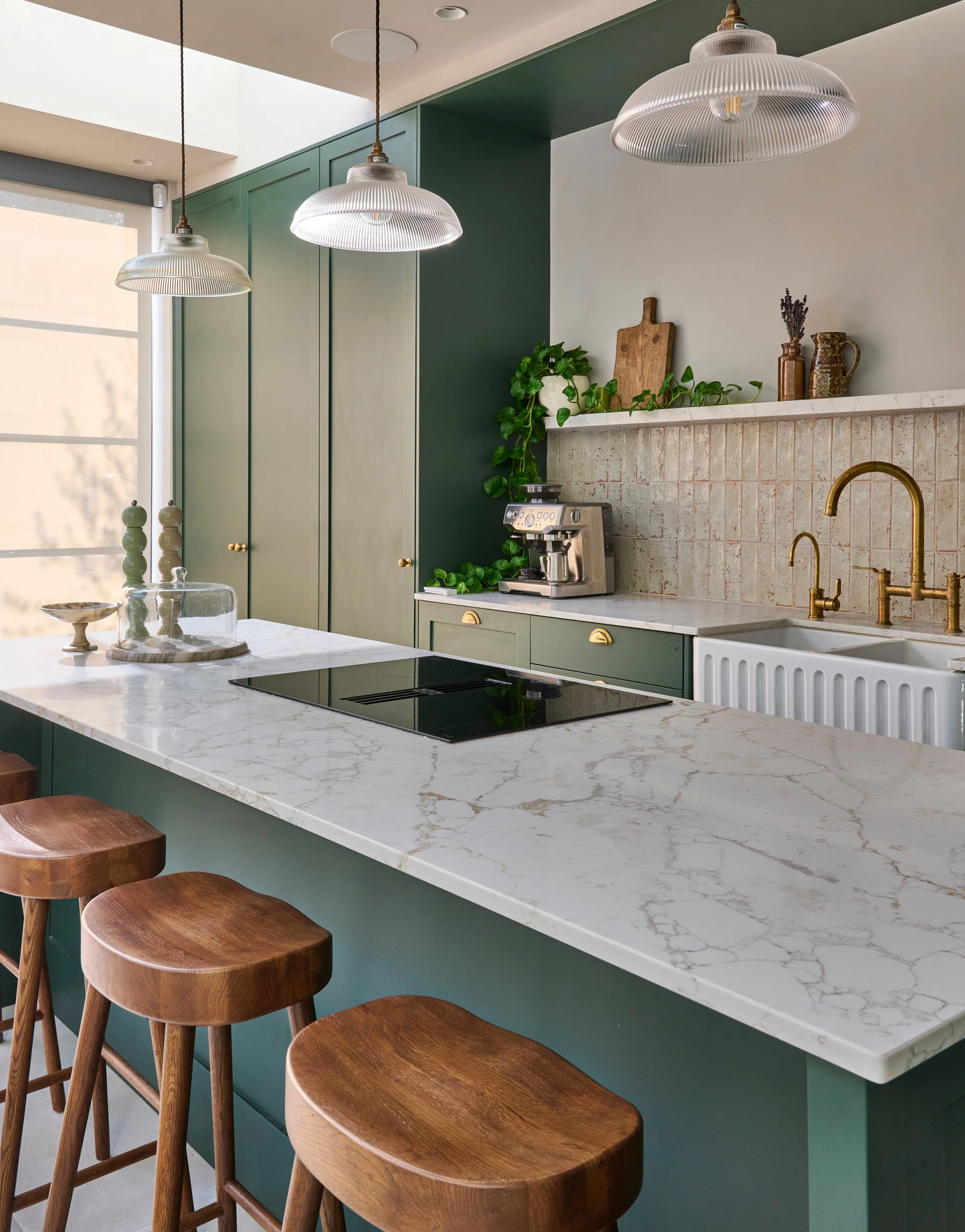 Modern kitchen with green cabinets, marble countertop, brass fixtures, and wooden stools.